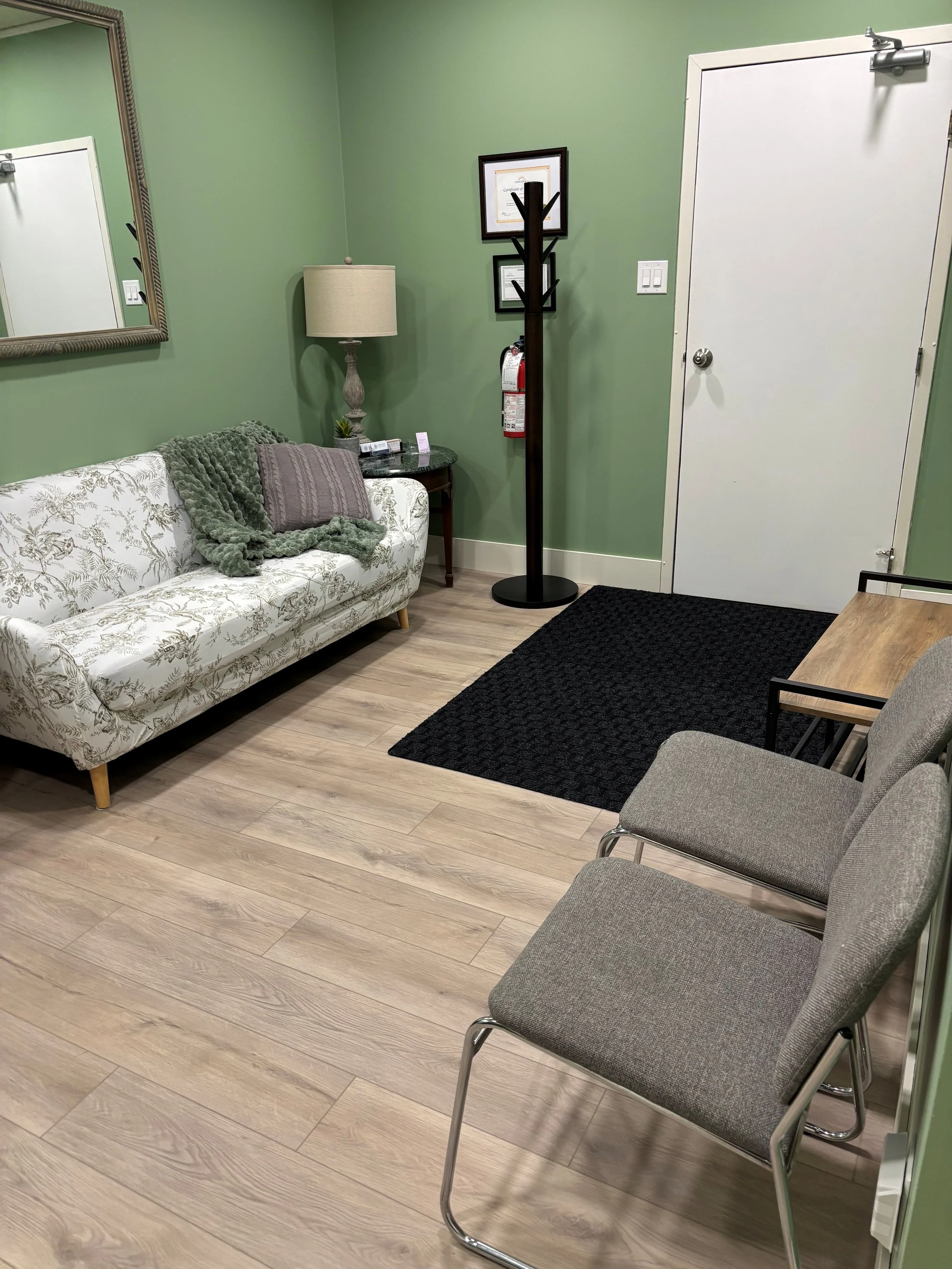 A warm and inviting waiting room with a soft and cozy floral-patterned white couch, a gray fabric chair, a black doormat, a wooden table, a side table with a lamp, a green wall, framed certificates, a coat rack, and a door in a therapy office.
