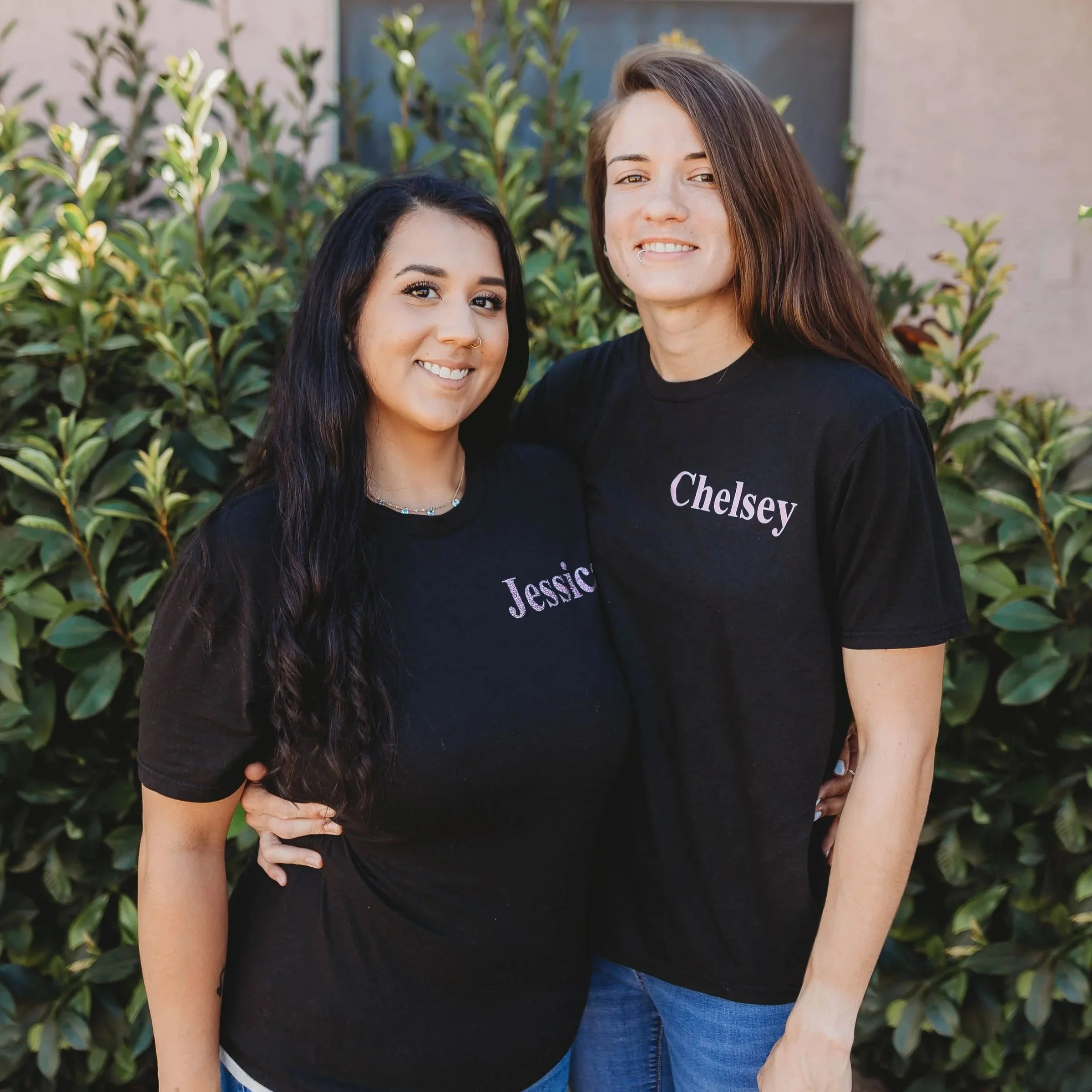 Two young women standing outdoors among green bushes, smiling and close together. One has long dark hair, light skin, and wearing a black shirt with her right hand on her hip. The other has long brown hair, light skin with freckles, and is wearing a black shirt