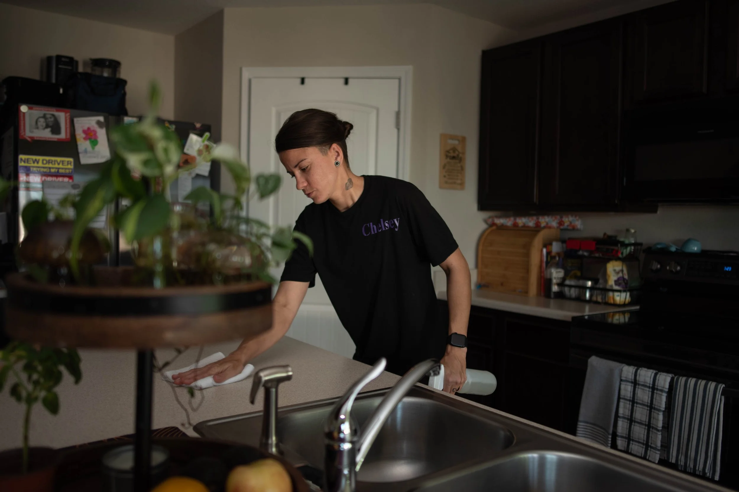 A woman with dark hair in a bun, wearing a black t-shirt with the name Chelsey, is cleaning the kitchen counter in a kitchen with dark cabinets and a white door.