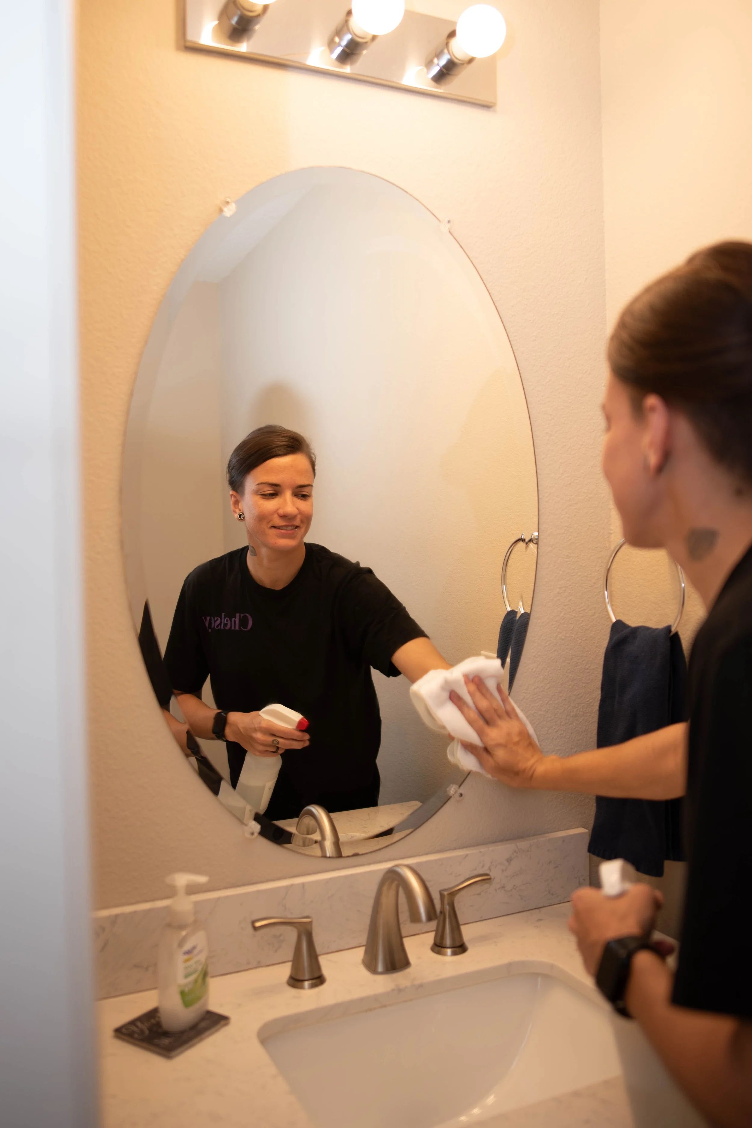 A woman with dark hair in a bun is standing in front of a bathroom mirror. She is holding a cloth in one hand and a spray bottle in the other, smiling while looking at her reflection. The bathroom has beige walls, a marble countertop, a sink, a soap dispenser, and a light fixture with four bulbs above the mirror.