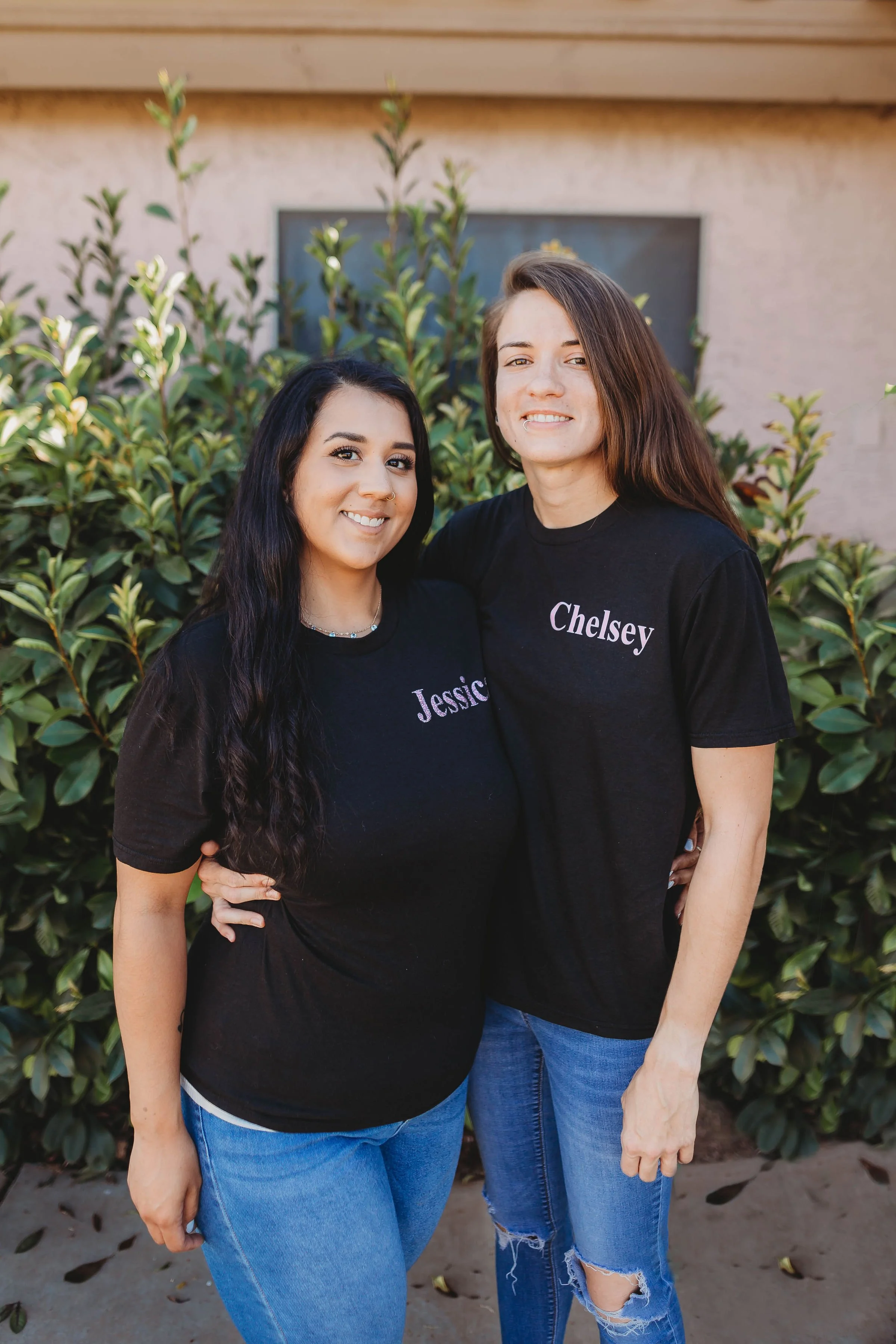 Two women standing outdoors in front of bushes, wearing black T-shirts with names 'Jessica' and 'Chelsey'.
