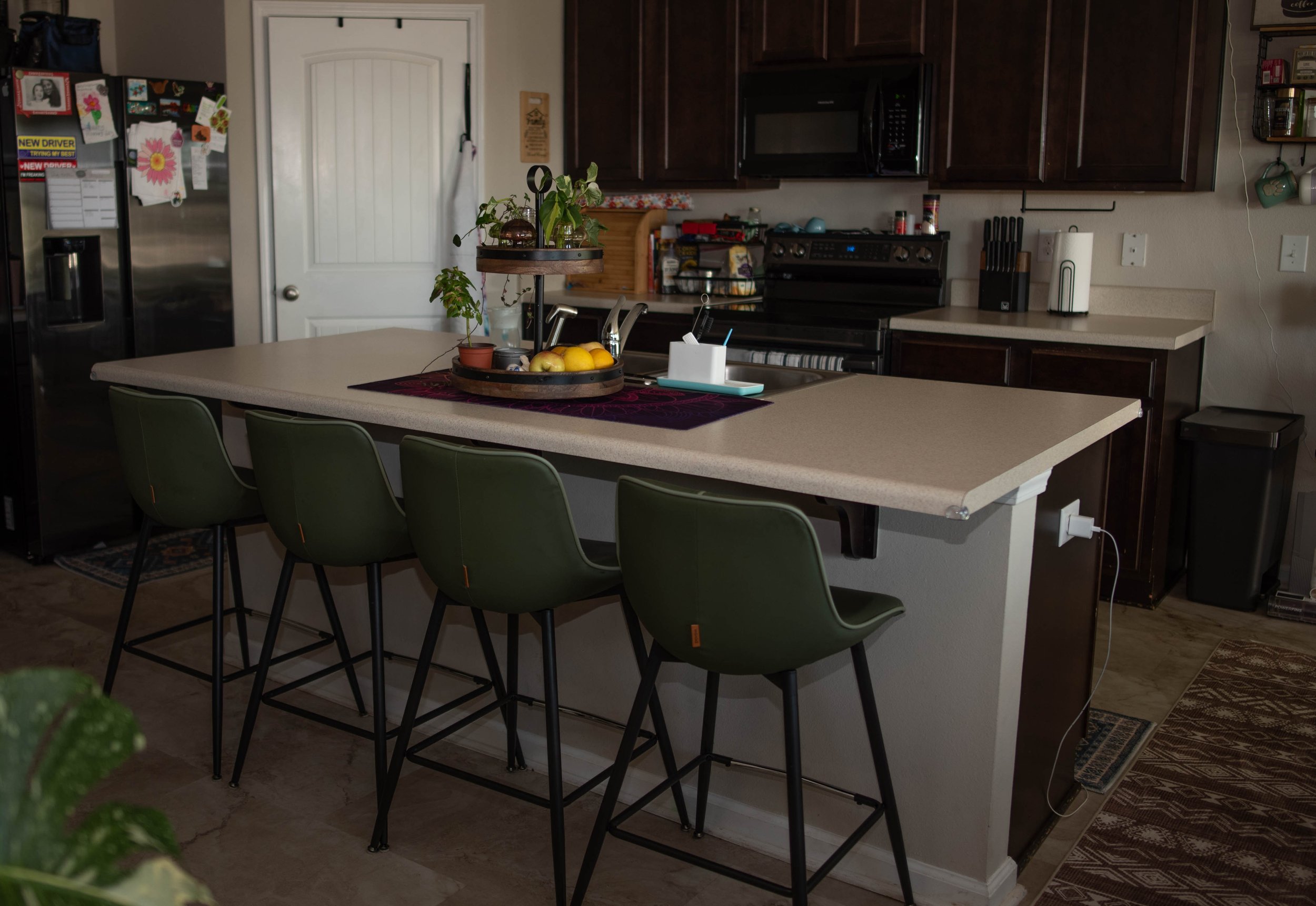 A kitchen with dark wooden cabinets, a microwave above the stove, a stainless steel refrigerator covered with magnets and notes, a countertop with a cutting board, a paper towel holder, and a fruit bowl, and a breakfast bar with green chairs.
