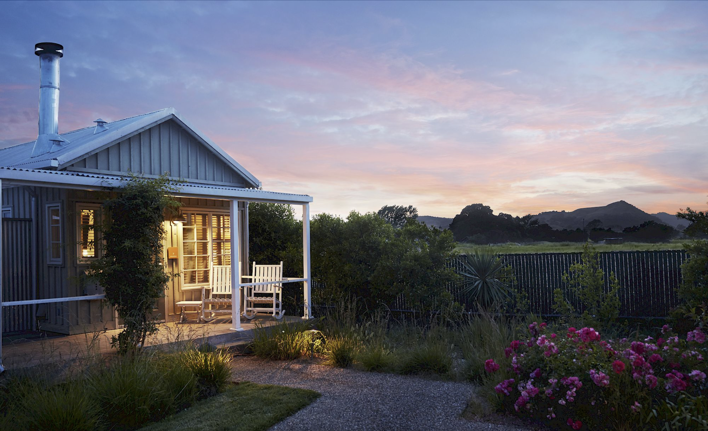 A cozy house with a porch and rocking chairs at sunset, surrounded by greenery and pink flowers, with mountains in the background.