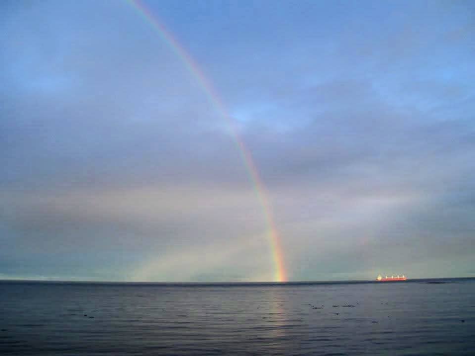 Rainbow over still water with patches of blue sky peaking through the clouds and cargo ship in background.