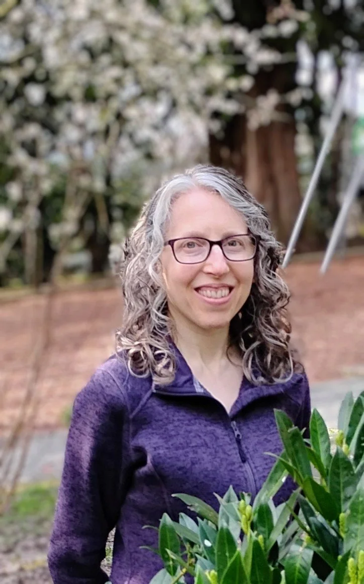 A woman with curly gray hair and glasses smiling outdoors, wearing a purple zip-up jacket, and standing with blossoming trees in the background.