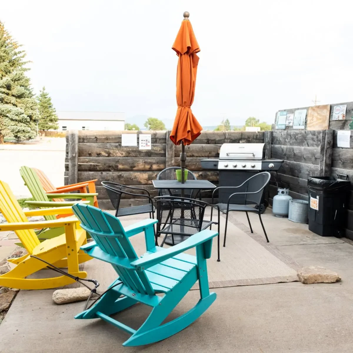 Outdoor seating area with colorful Adirondack chairs, a glass table with a large orange umbrella, a grill, and a fenced backdrop.