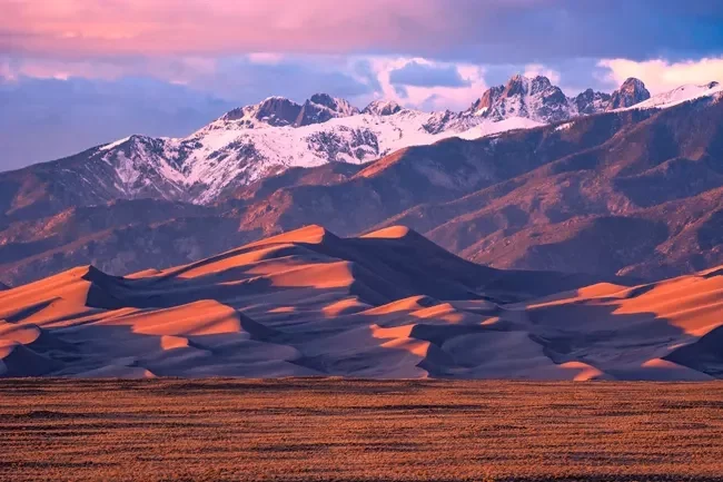 Sand dunes in the foreground with snow-capped mountains in the background during sunset or sunrise.