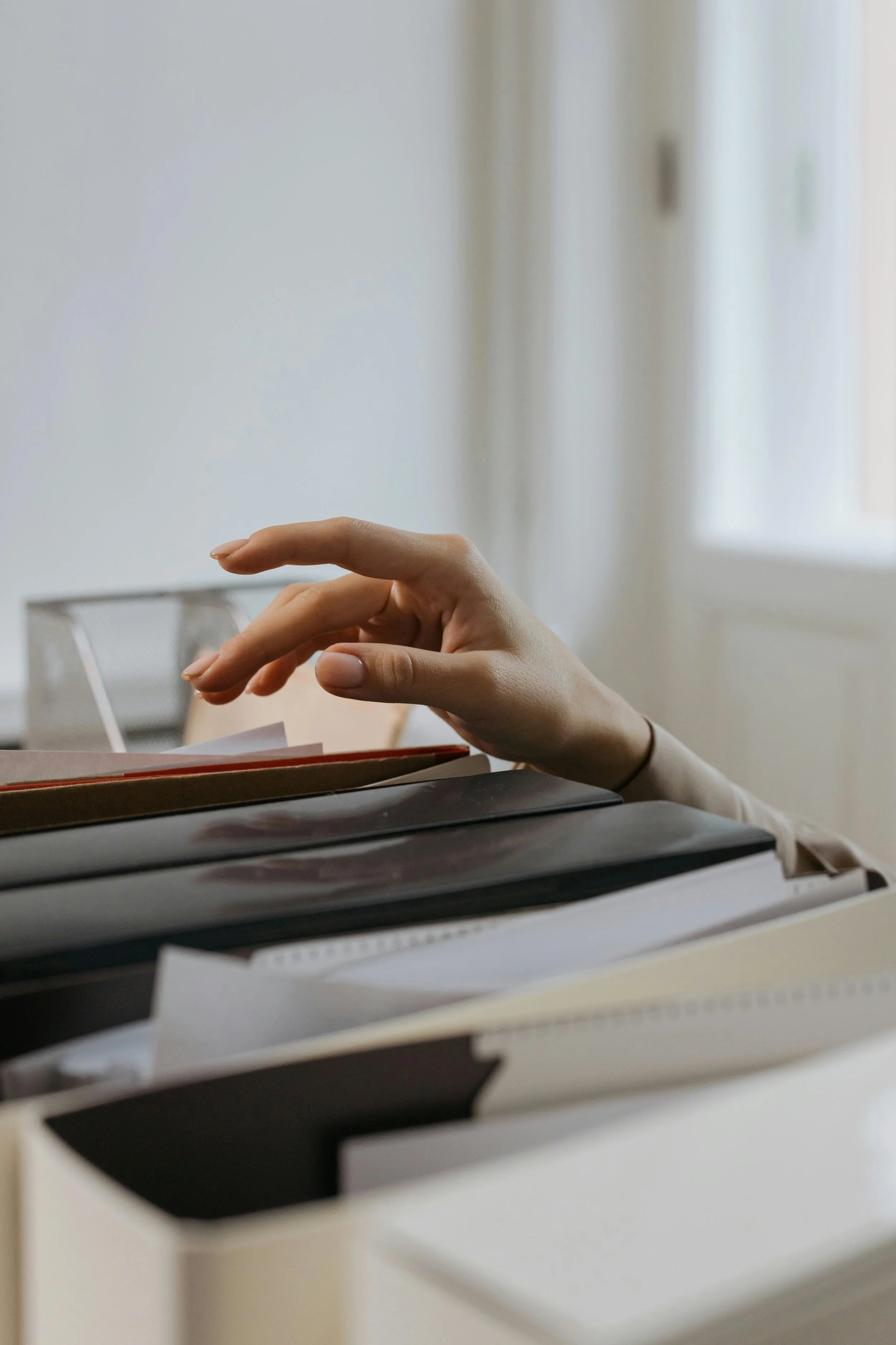 A person's hand reaching over a stack of files and documents on a desk in a bright room.