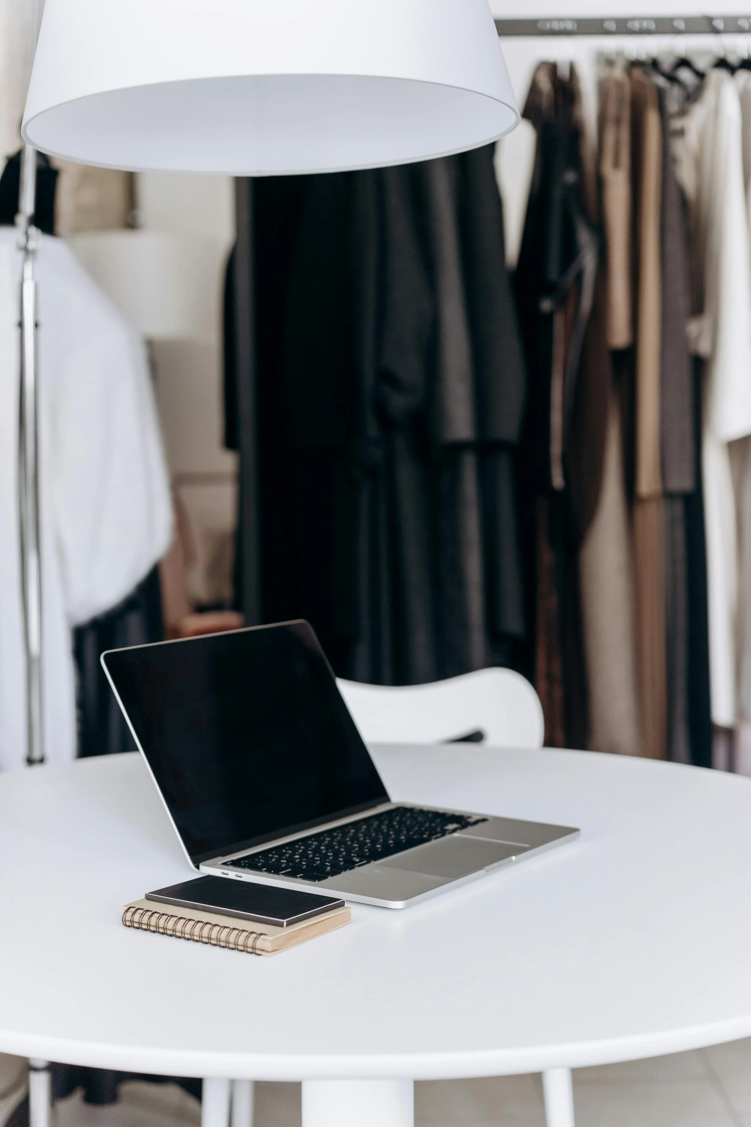 Open laptop, smartphone, and spiral notebook on a white table in a room with clothing racks in the background.