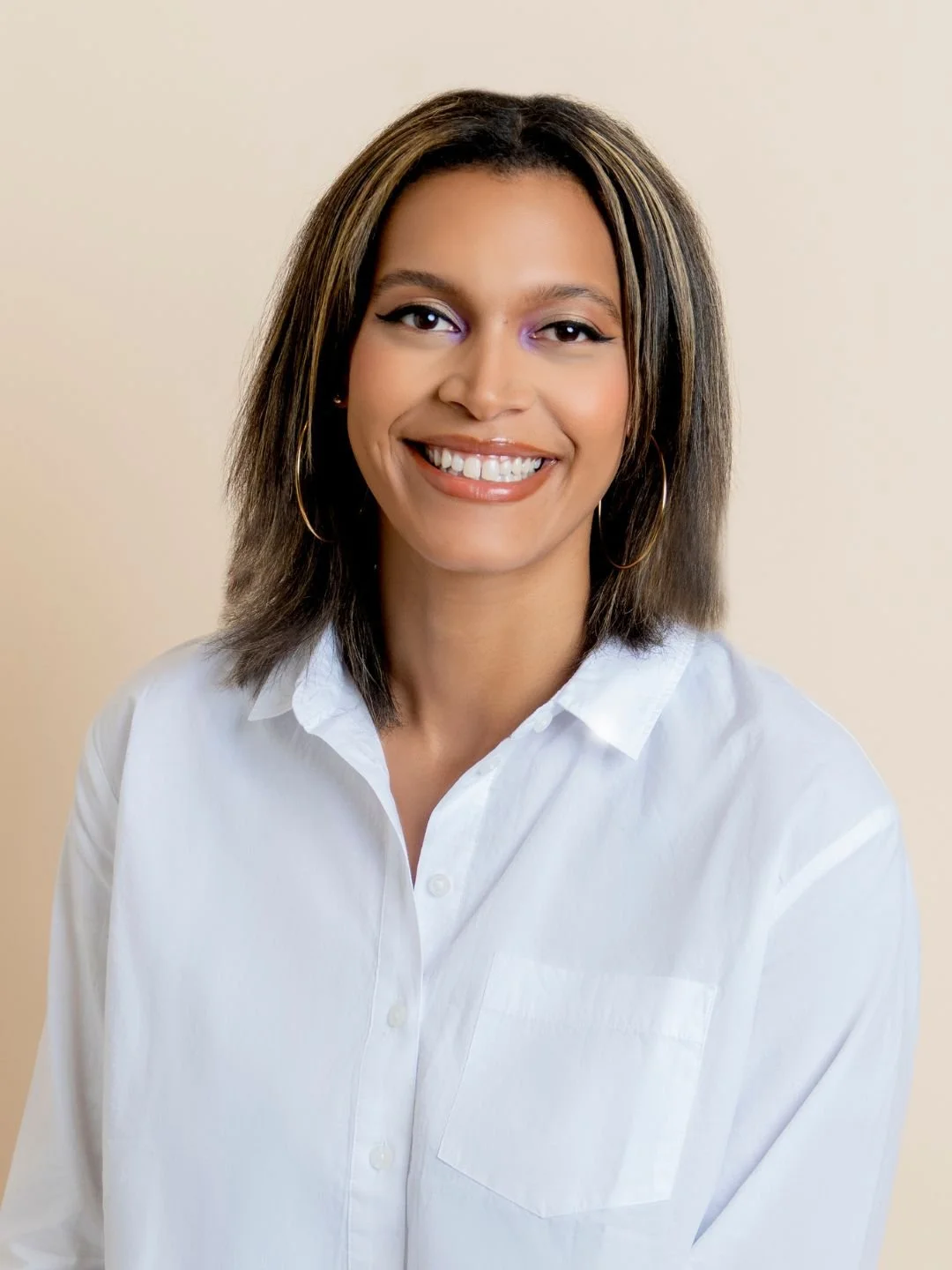 Smiling woman with shoulder-length brown hair, wearing a white button-up shirt and hoop earrings, posing against a neutral background.
