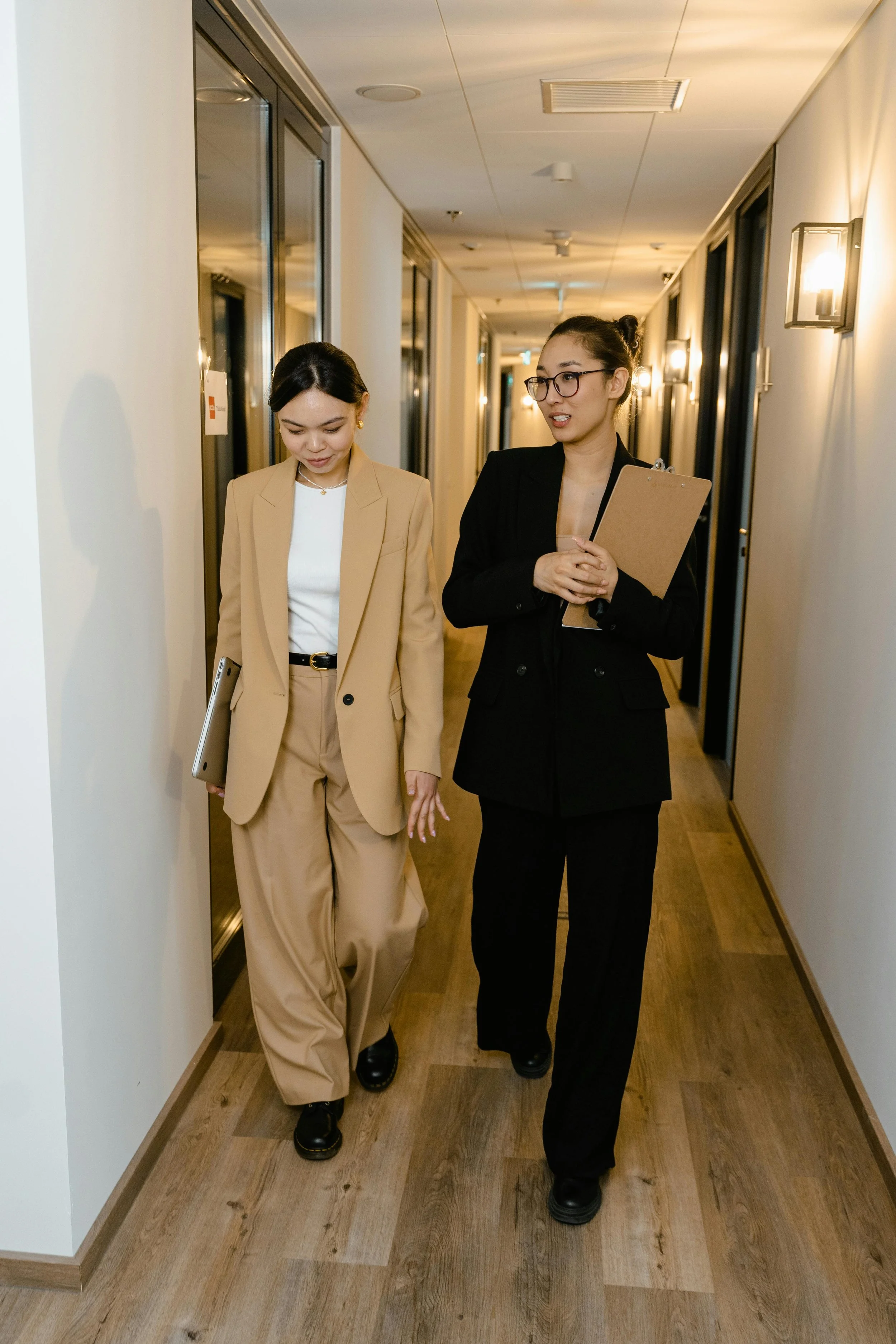 Two women walking down a hallway, engaged in conversation. One is dressed in a beige suit and holding a laptop, the other in a black suit holding a clipboard.