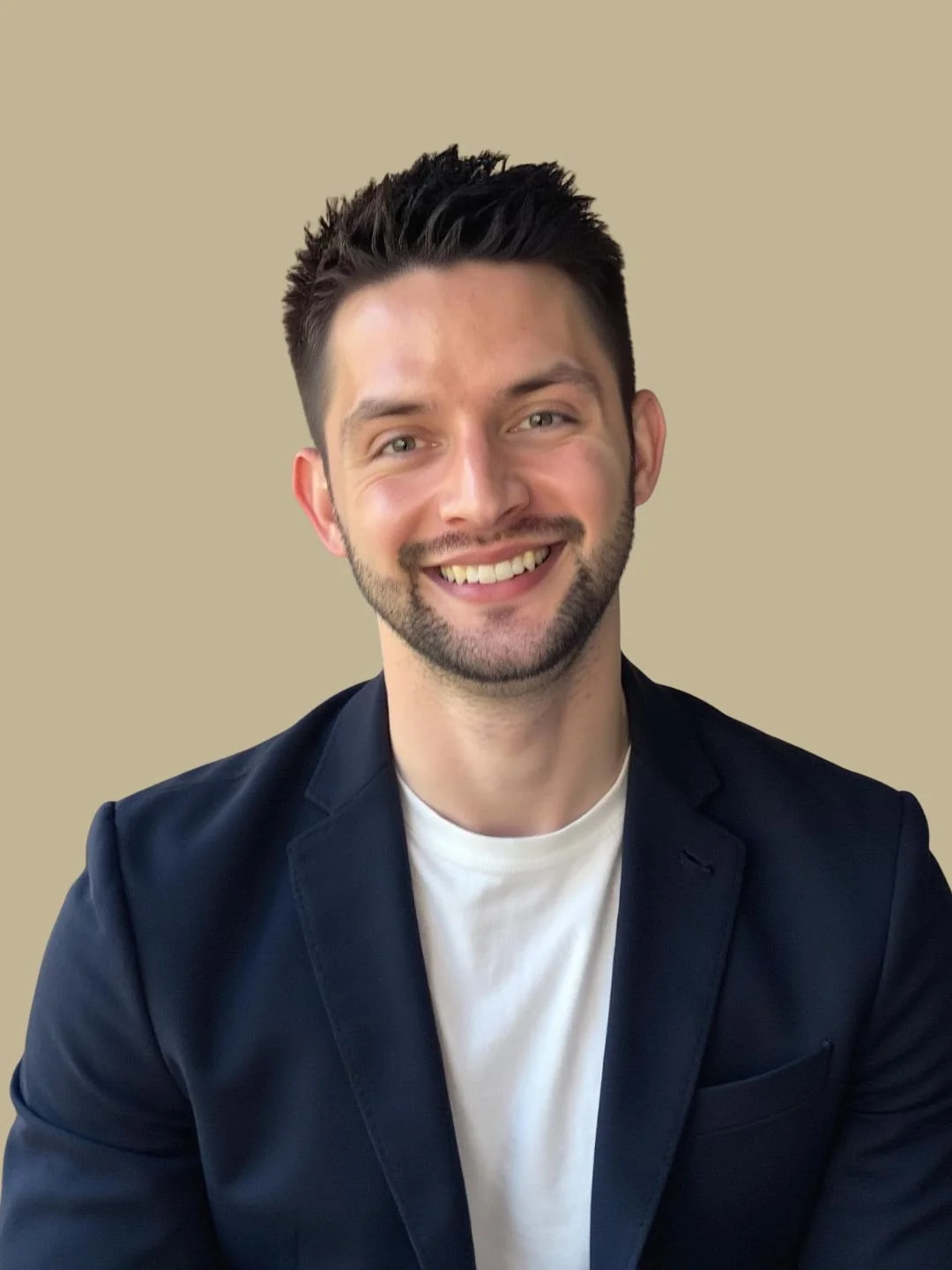 Portrait of a young man with dark hair and a beard, smiling, wearing a white t-shirt and a dark blazer, against a beige background.