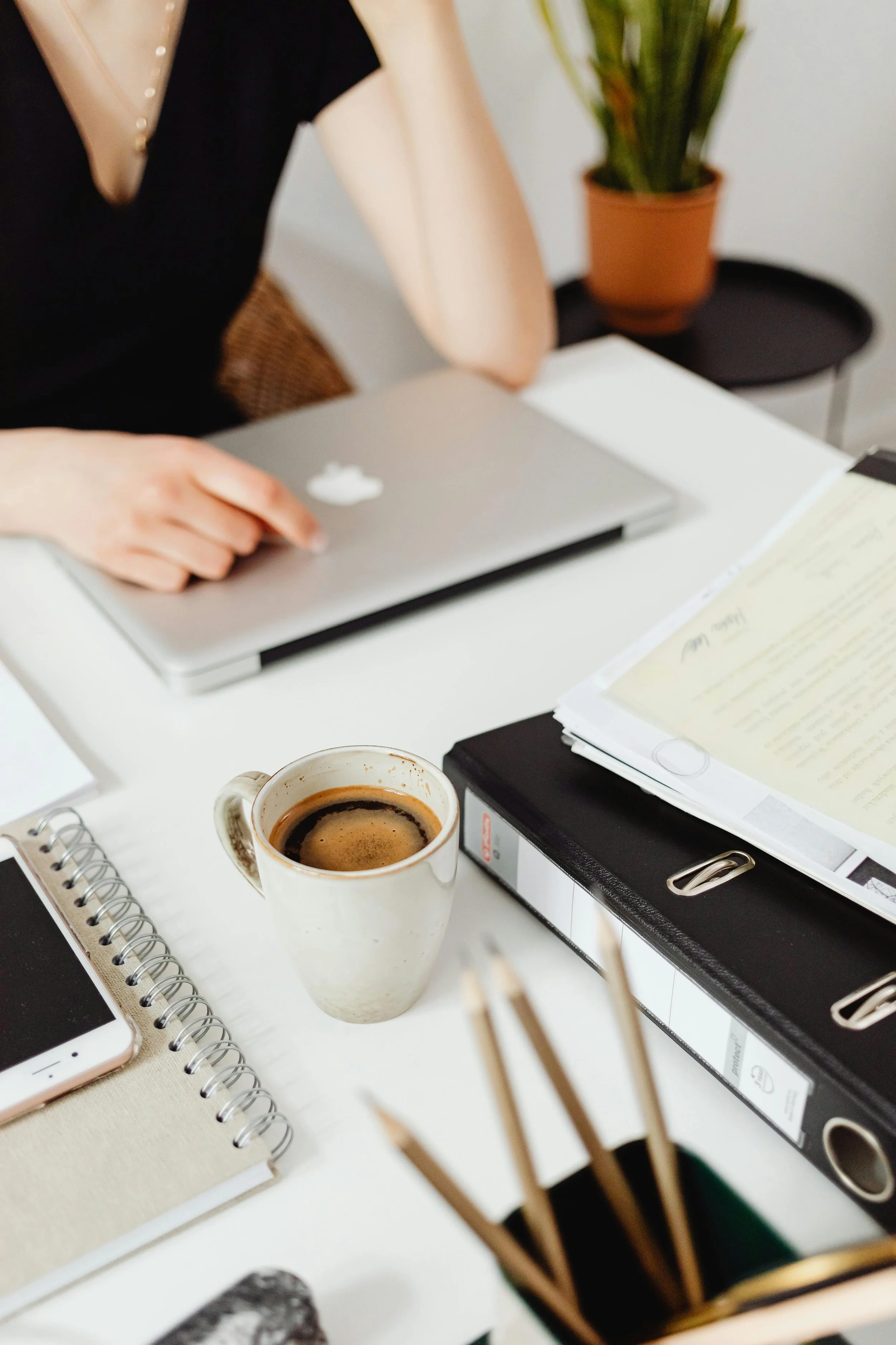 A cluttered white desk with a cup of coffee, a smartphone, notebooks, a black binder, and a laptop with a person working in the background.