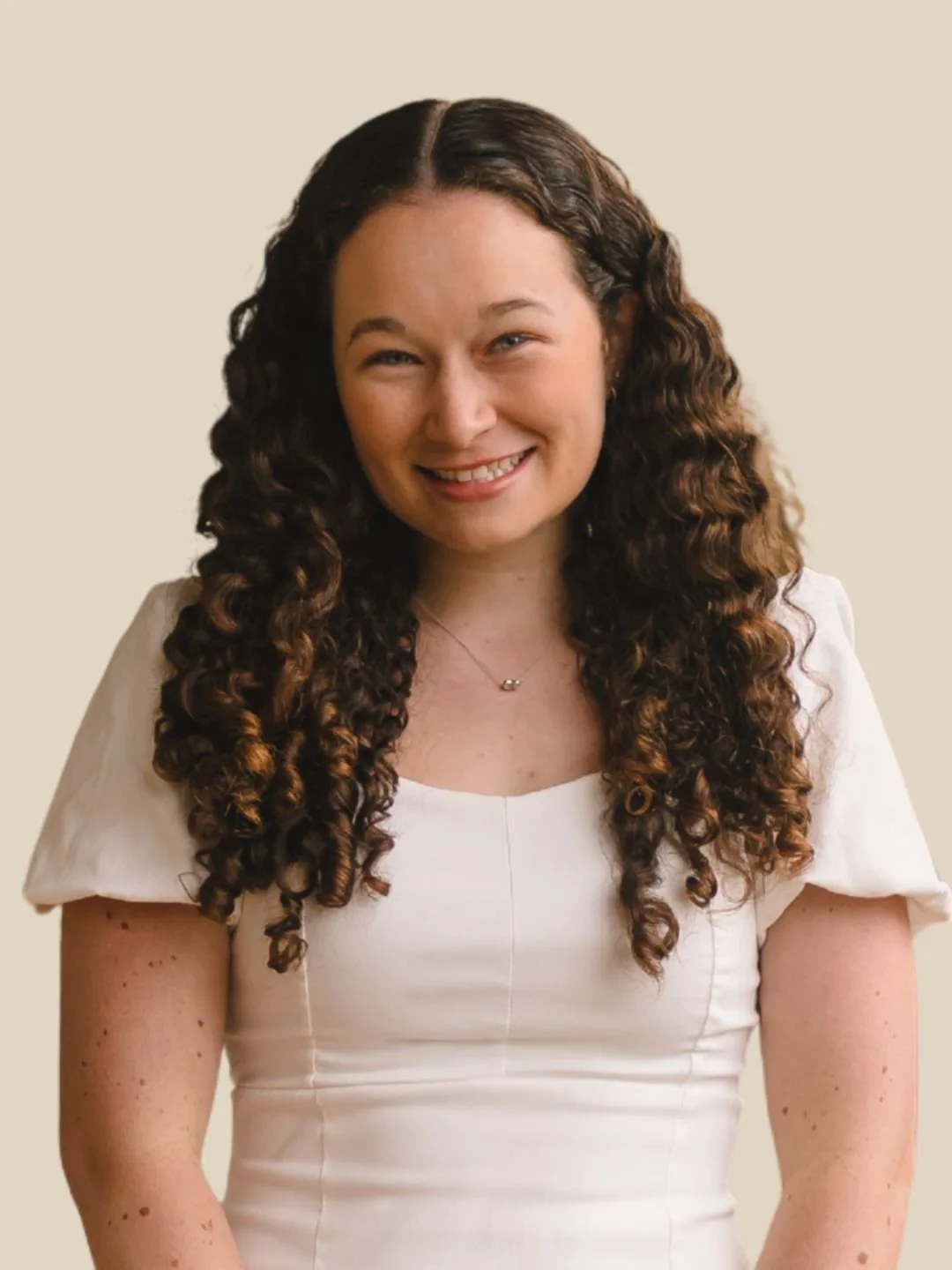A woman with long, dark curly hair smiling, wearing a white top and a delicate necklace, against a light beige background.