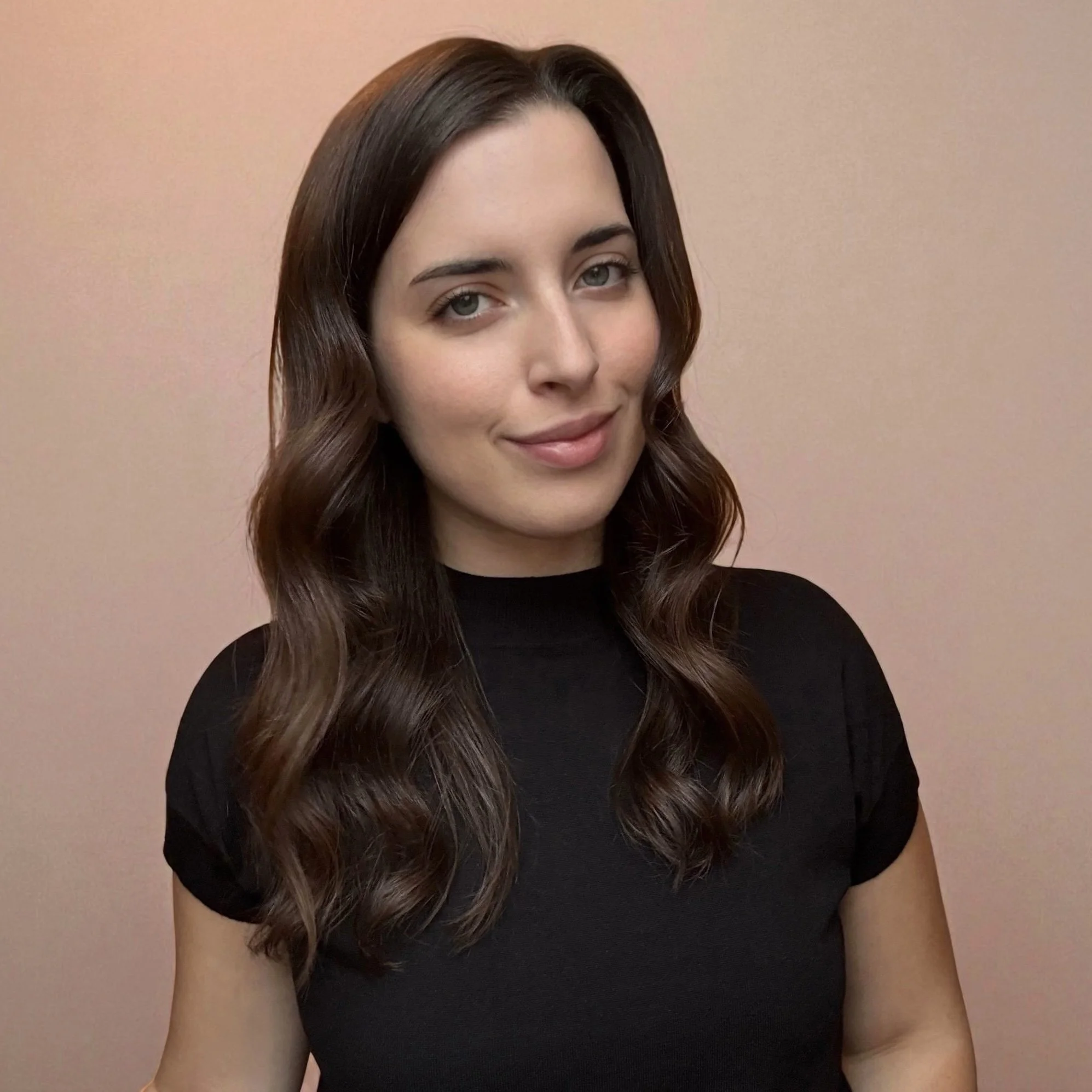 A woman with long, wavy brown hair wearing a black top, smiling softly against a plain, light-colored background.
