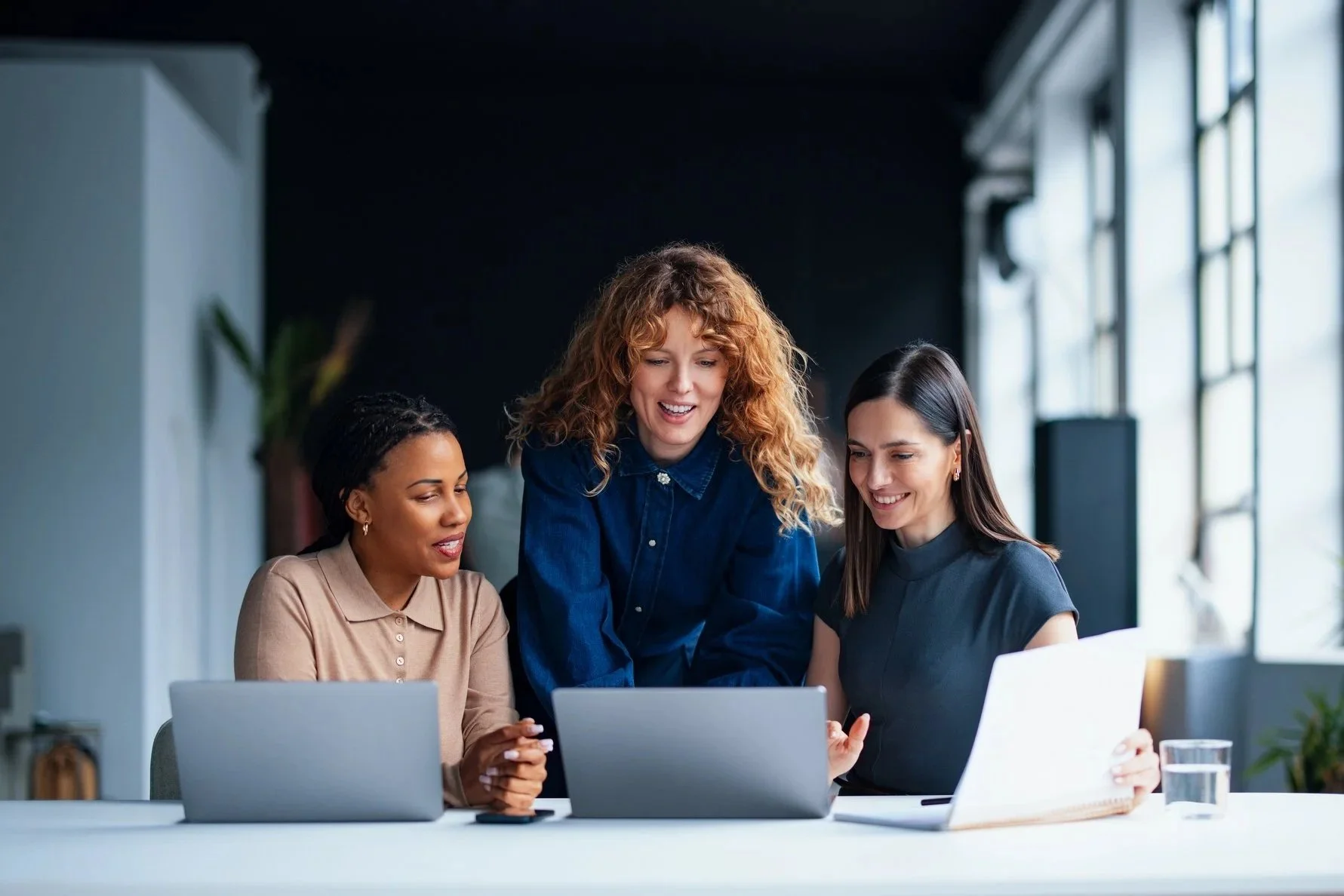 Three women working together at a table with laptops in a modern office, collaborating and smiling