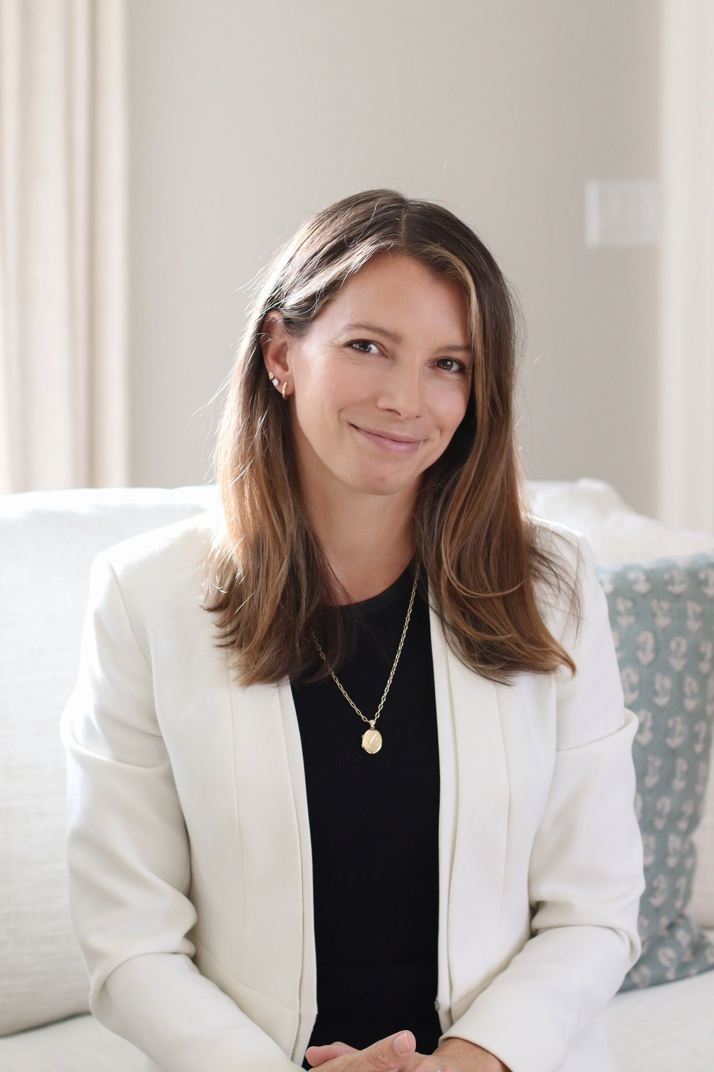 A woman with brown hair and a slight smile, wearing a white blazer and a black top, sitting on a light-colored couch with an anchor-patterned pillow in the background.