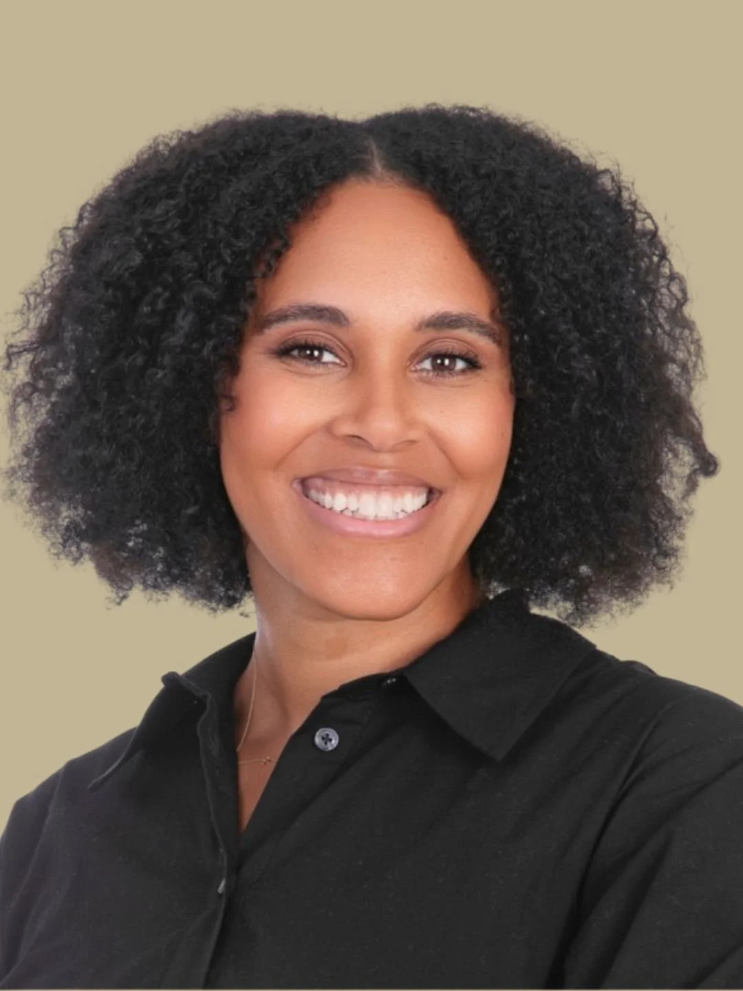 A woman with dark, curly hair and brown skin smiling, wearing a black shirt against a neutral background.