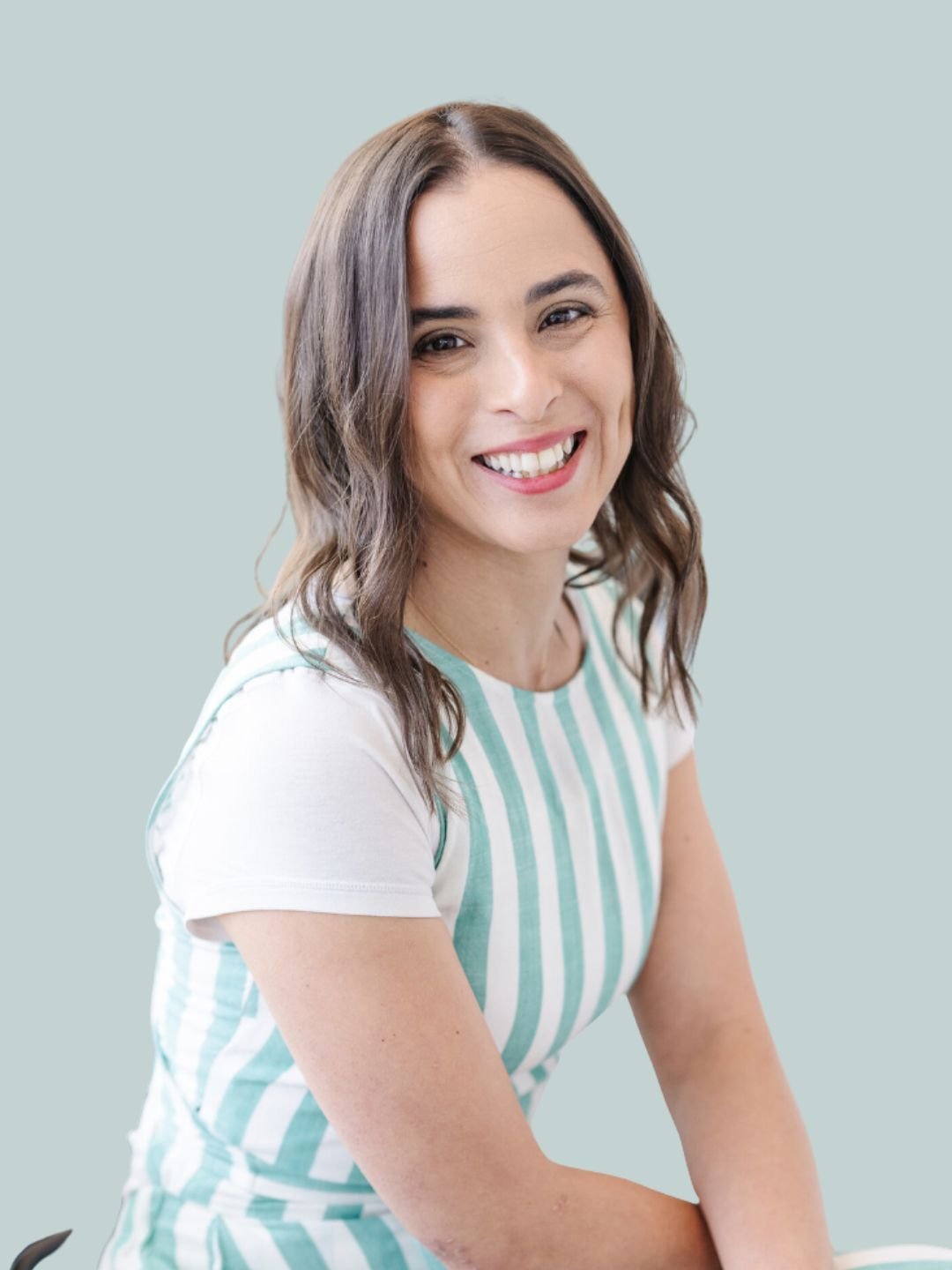 A young woman with shoulder-length brown hair smiling, wearing a white and light blue striped t-shirt, against a plain light grey background.