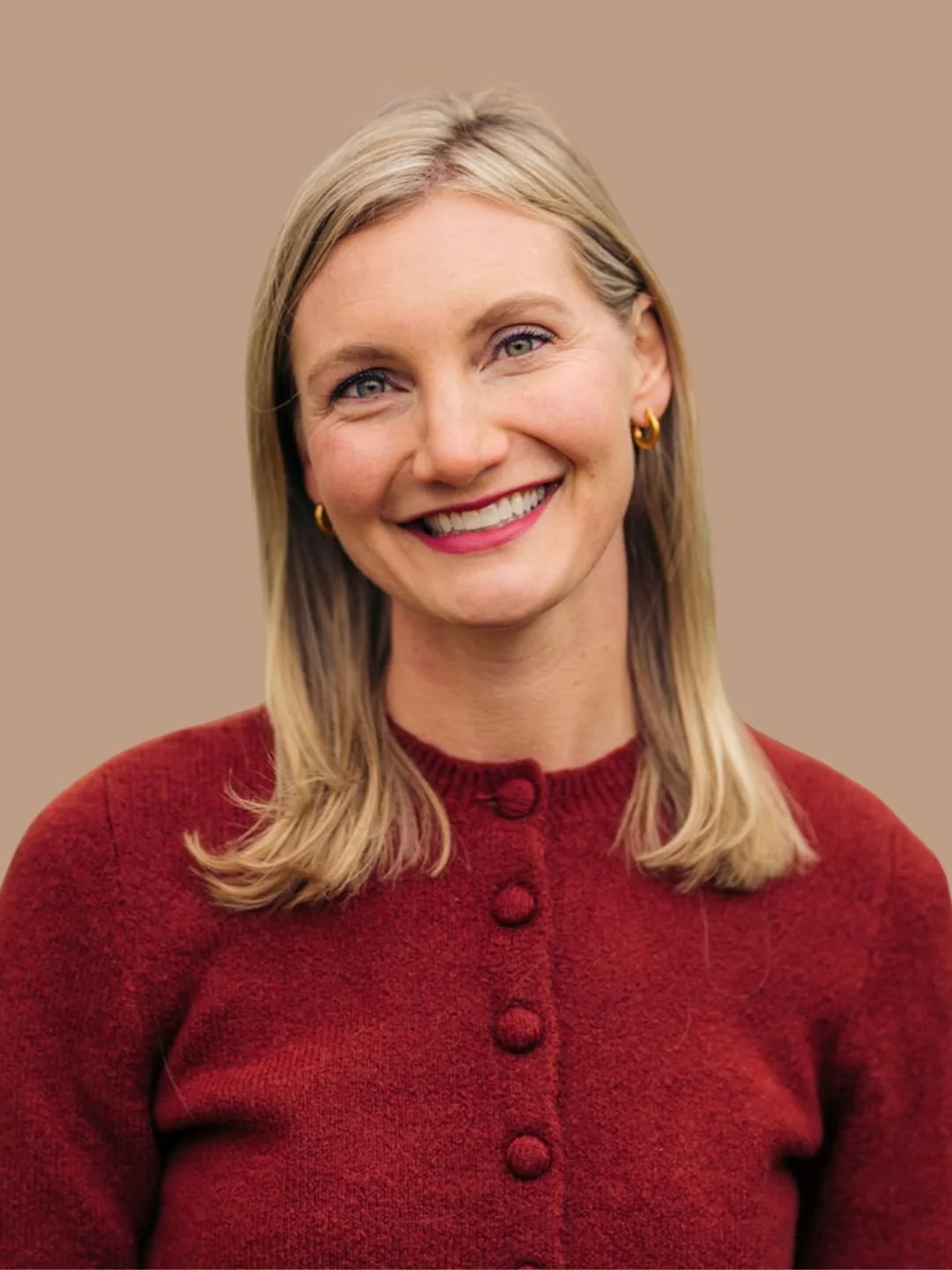 A smiling woman with blonde hair, wearing gold hoop earrings and a red sweater with buttons, standing against a neutral background.