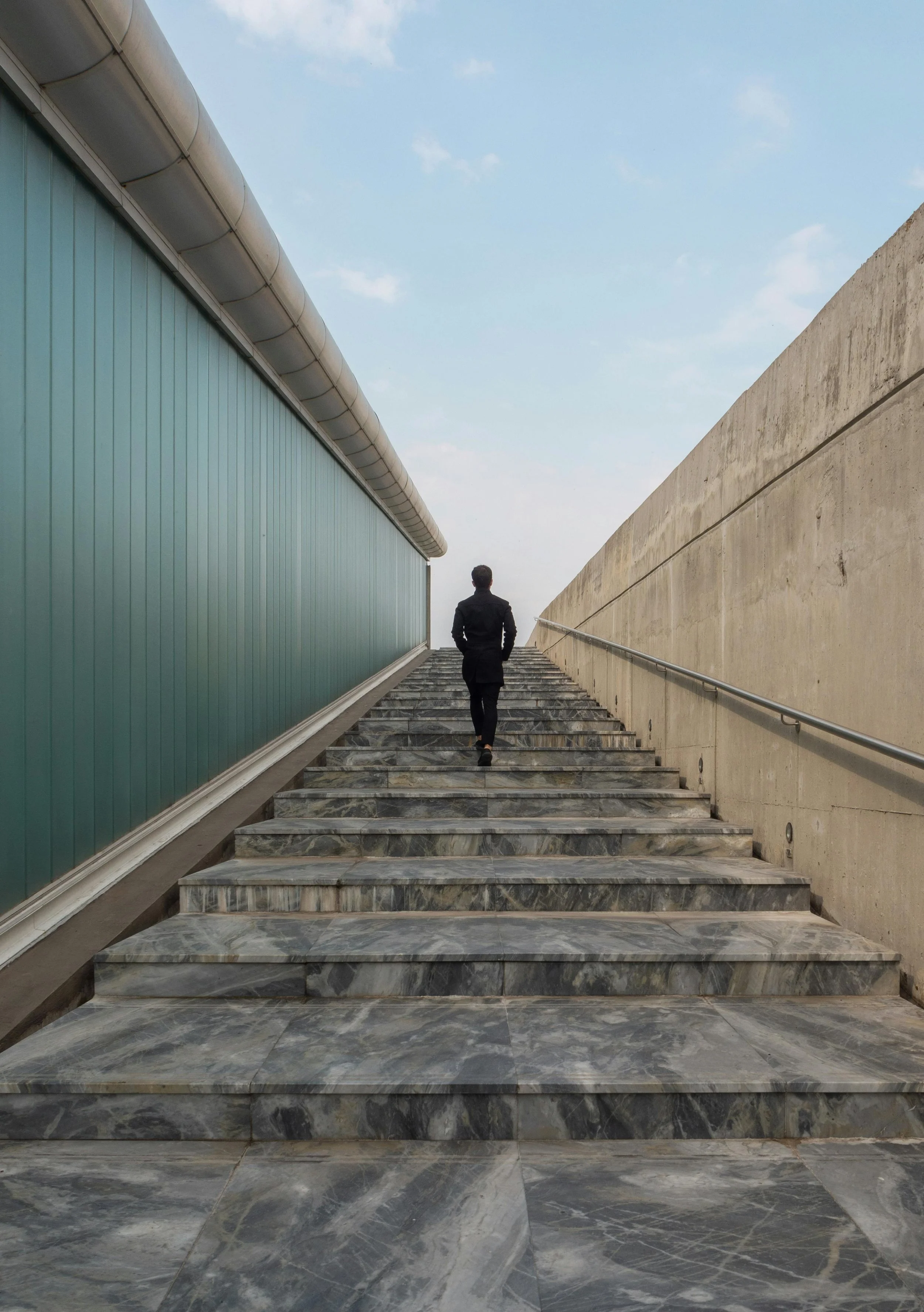 A man in black walking up a marble staircase with a green building on the left and a concrete wall on the right, under a partly cloudy sky.