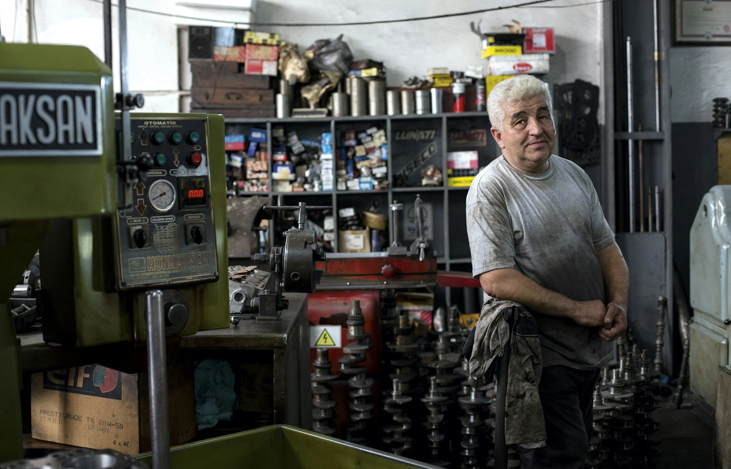 A man with gray hair and a gray shirt stands in a cluttered workshop with shelves filled with tools, parts, and boxes.