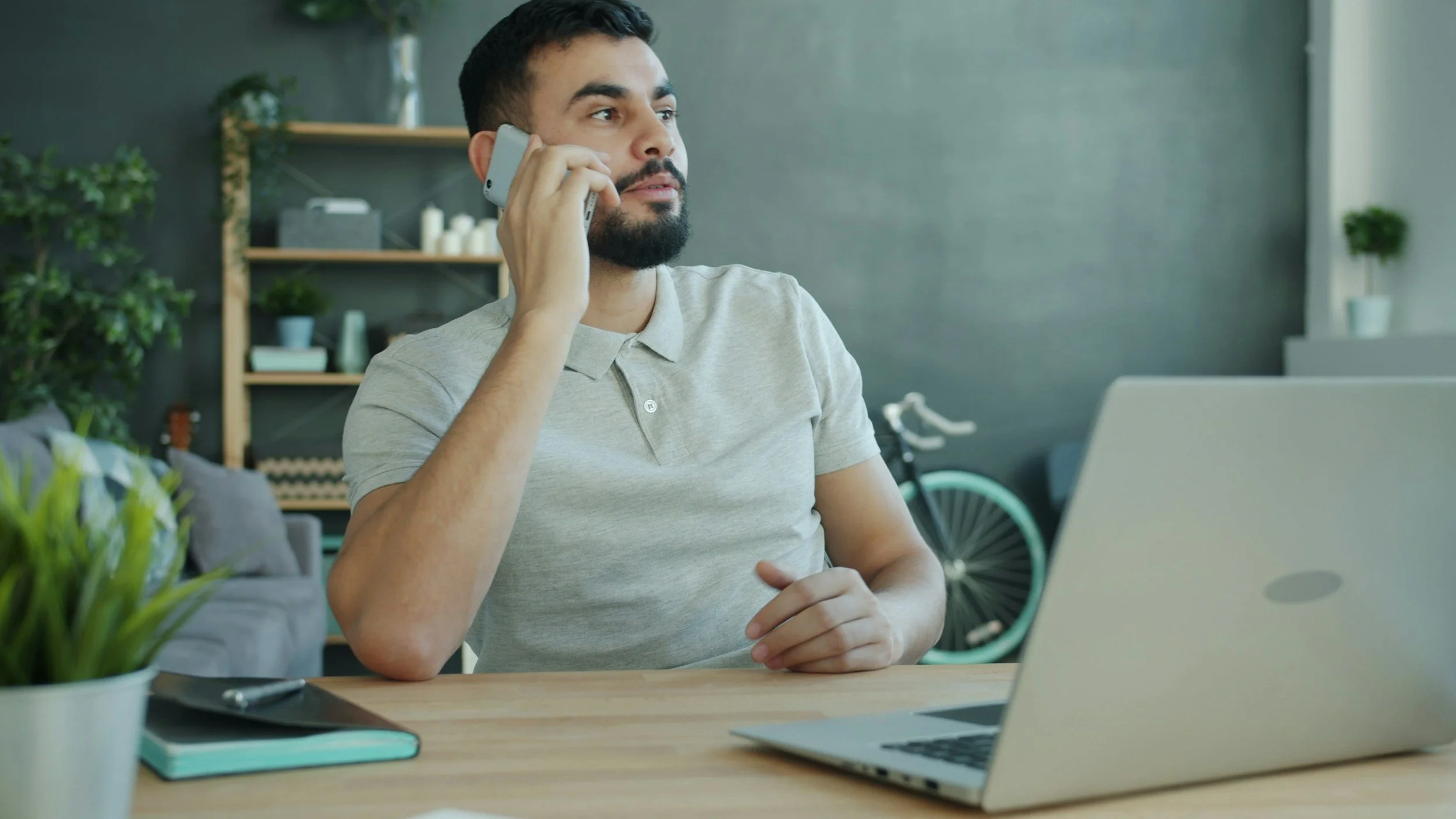 A man in a gray polo shirt sitting at a desk talking on a phone. There is a laptop, a notebook, and a pen on the desk, with a potted plant nearby. The background includes shelves with decorative items and a bicycle against the wall.