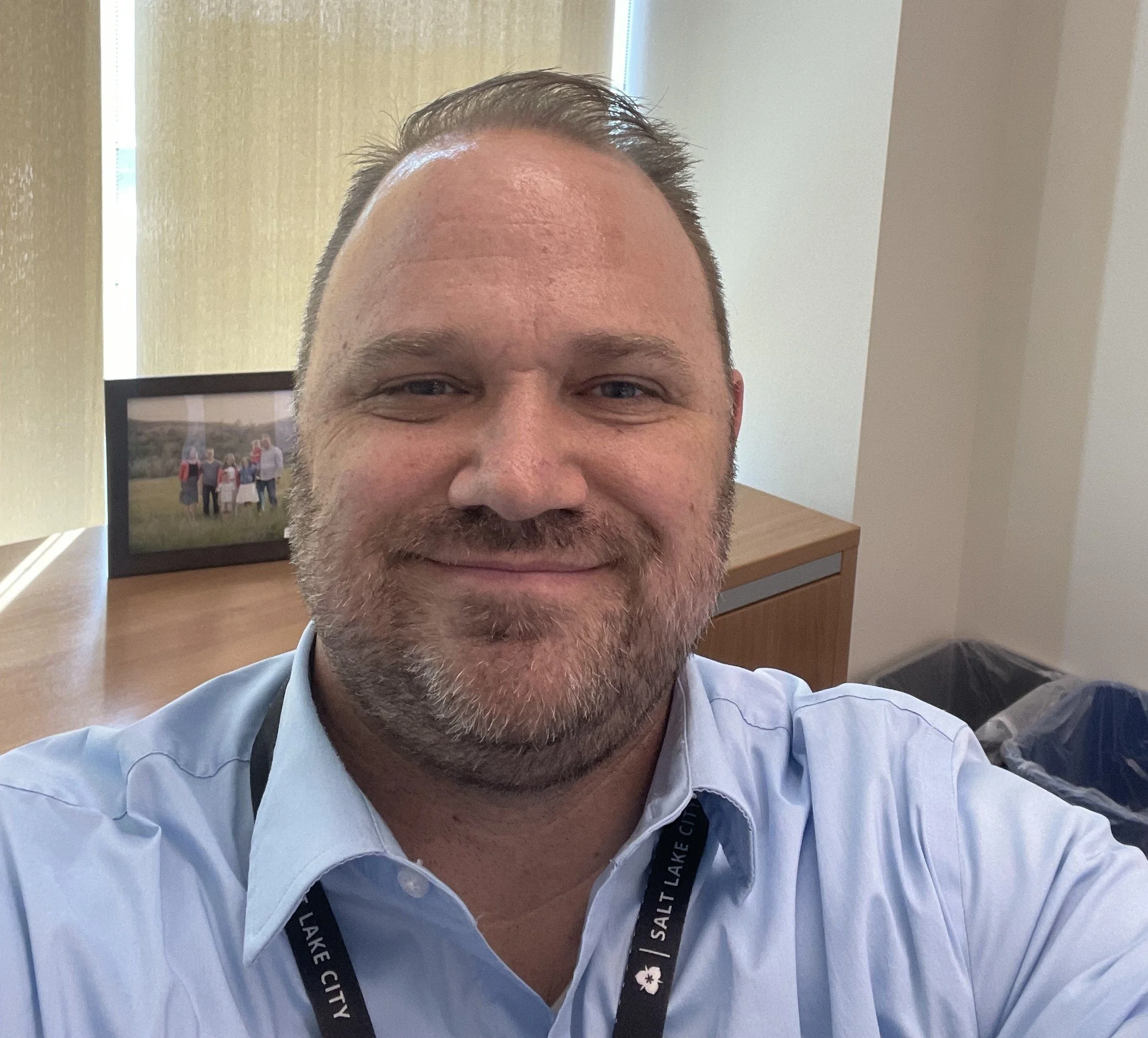 A man with short hair and a beard taking a selfie, wearing a light blue dress shirt and a lanyard that reads 'Salt Lake City.' Behind him, a framed photo of a group of people holding a flag and standing outdoors is visible, along with a wooden desk and a trash bin.
