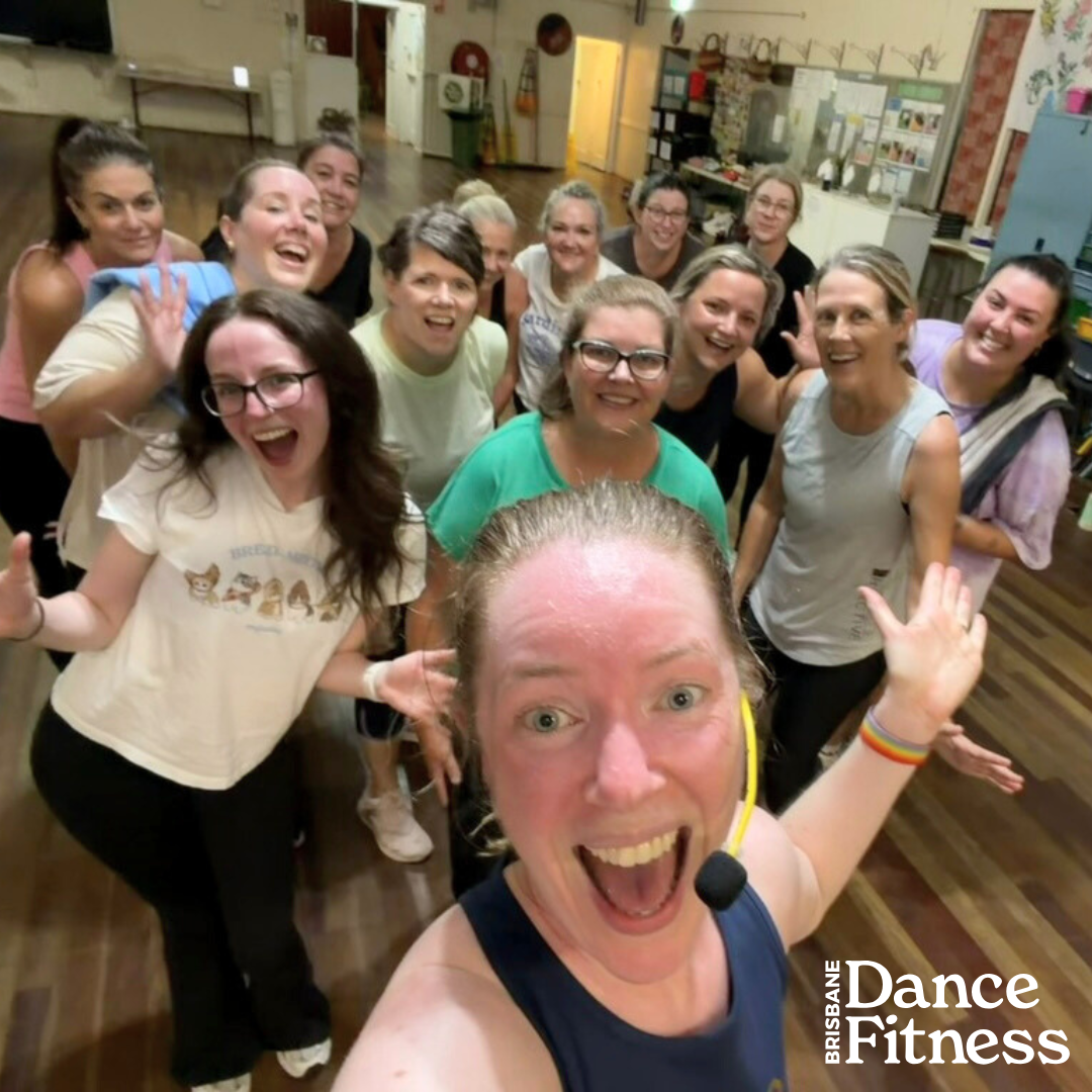 Group of women smiling and posing together in a dance fitness class, with the instructor taking a selfie at the front, in a studio with wooden floors and a decorated wall.