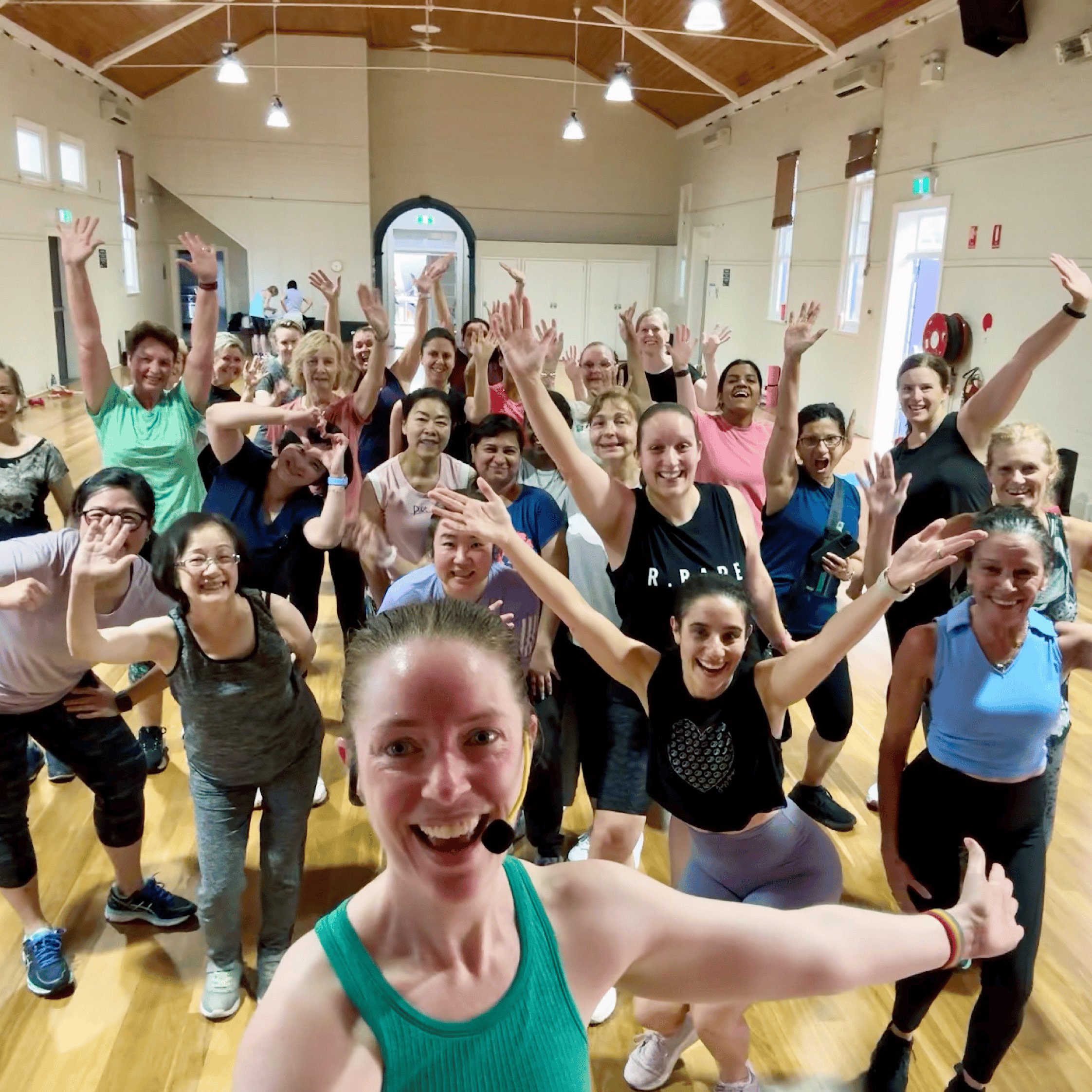 Group fitness class in a gym with smiling women and a female instructor taking a selfie in the foreground.