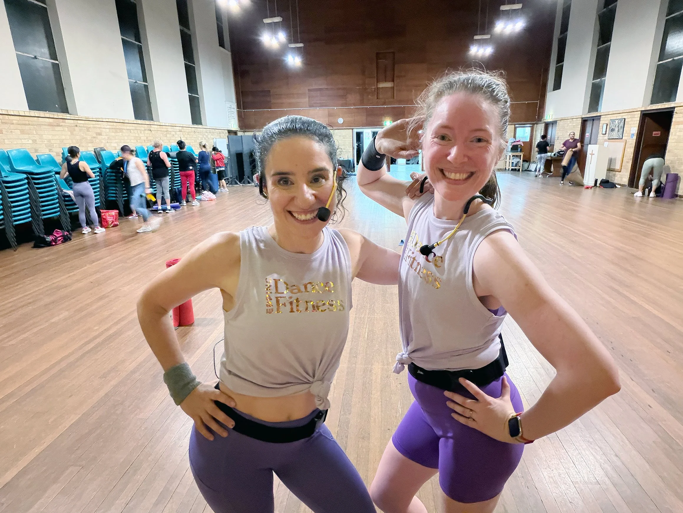 Two smiling women in workout clothes pose together in a dance fitness class studio, with others exercising in the background.
