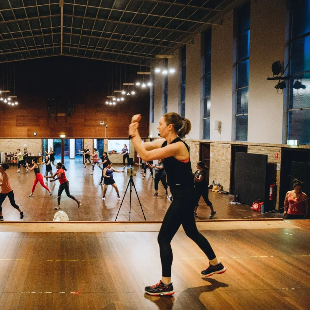 A dance or fitness class in a large studio with wooden floors and high ceiling. Multiple women are participating, following an instructor who is leading from the front. The instructor is a woman with a ponytail, wearing black athletic wear and sneakers, gesturing with her arms. The room has large windows and hanging lights.