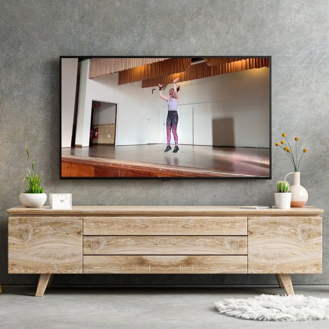 Living room with a wall-mounted flat-screen TV displaying a woman in workout clothes performing an exercise with a barbell. The TV is above a wooden console table decorated with a small clock, green plants, and a vase with orange flowers. Light-colored curtains are partially visible on the left. The wall behind is textured gray and the floor is light-colored with a small white rug in front of the console.