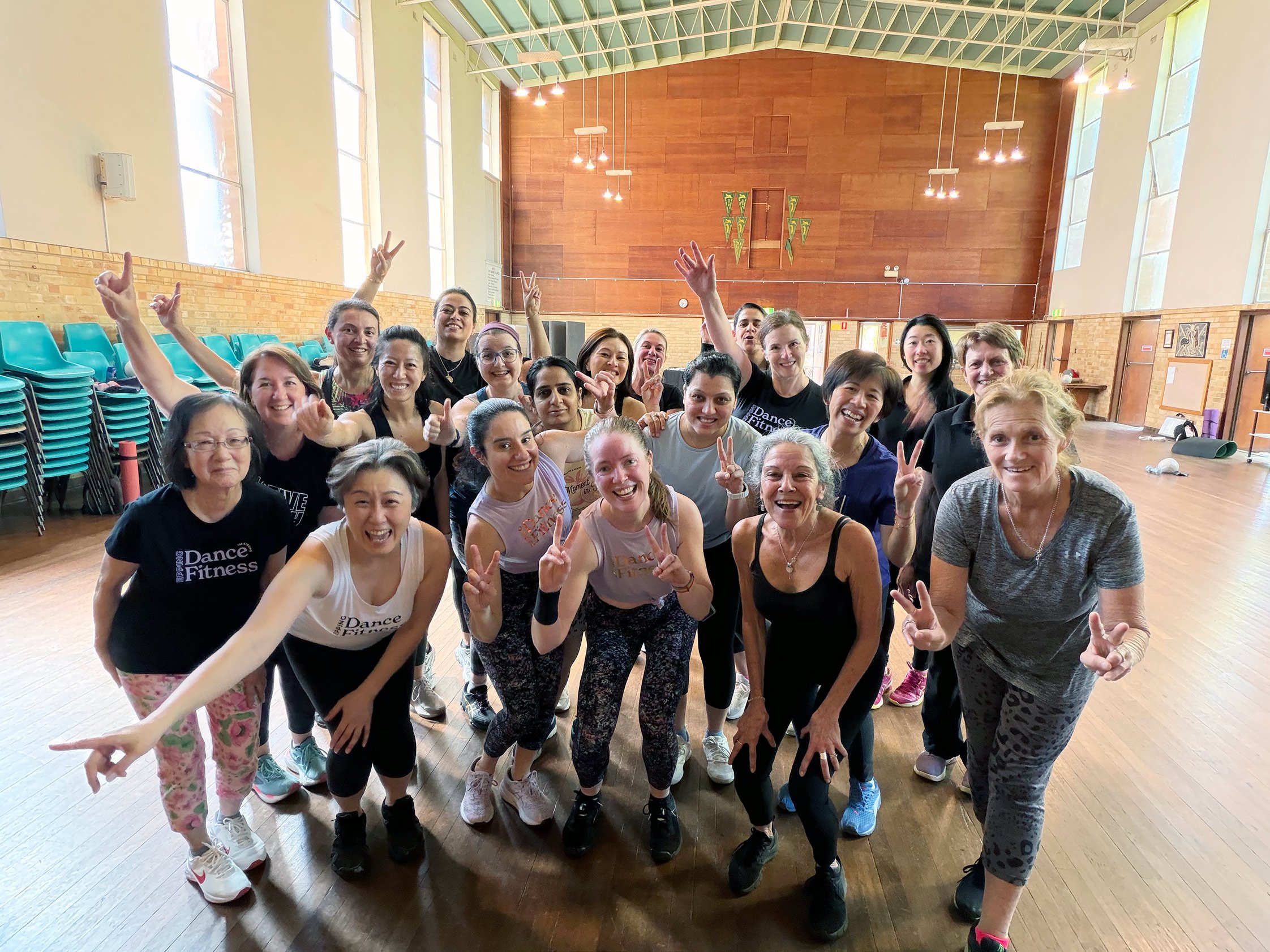 Group of women in athletic attire smiling and posing together in a gymnasium, some making peace signs, with friends and fitness instructors.