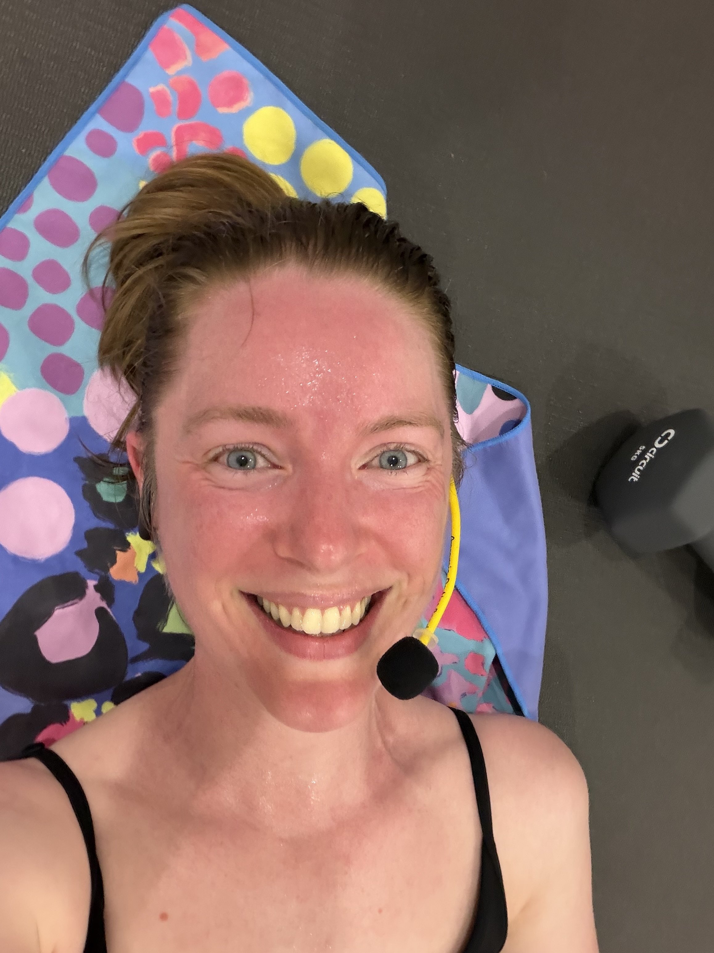 Instructor, Fiona, lying on a yoga mat. She is wearing a microphone, is smiling, and looks sweaty.