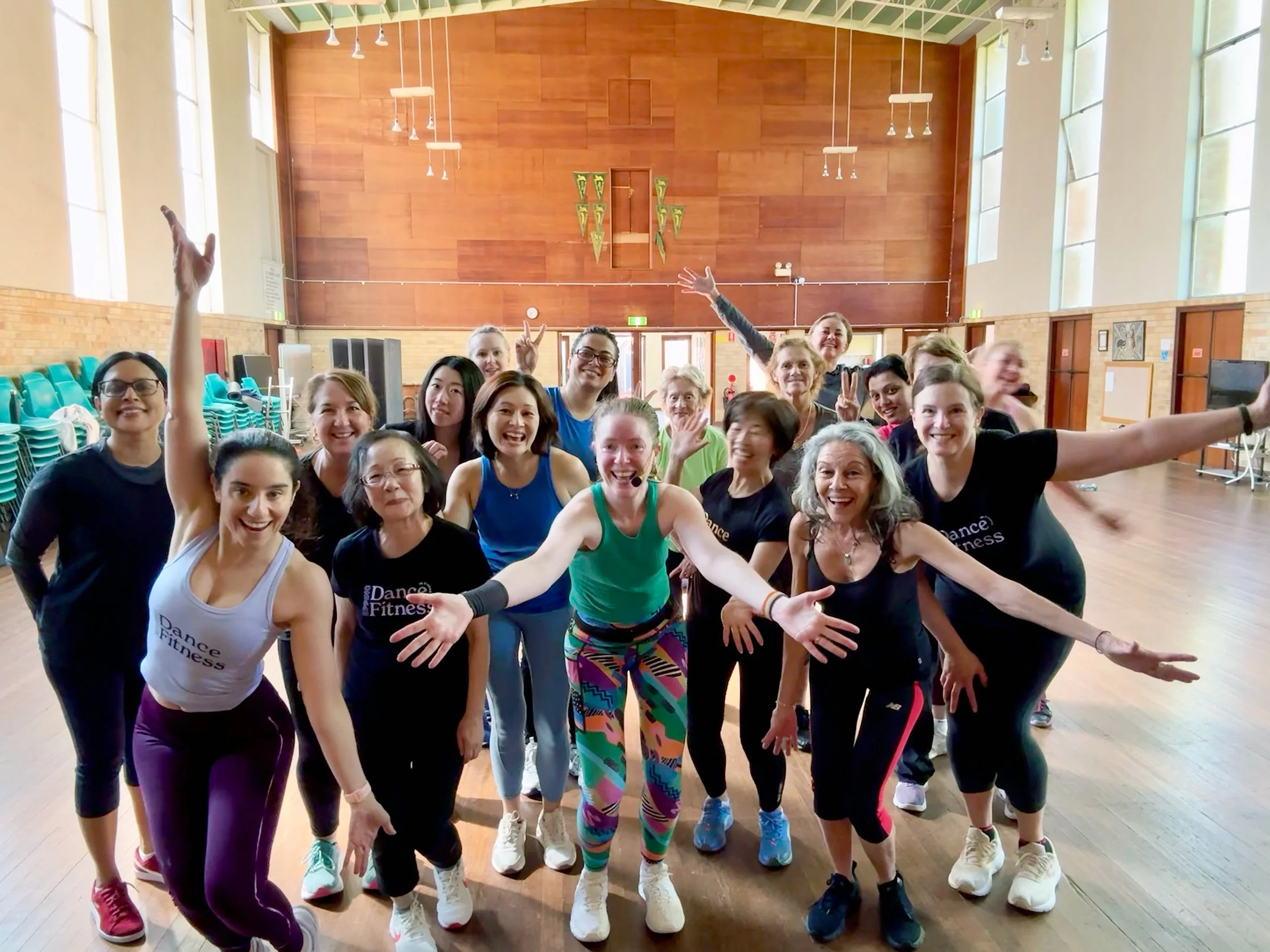 A group of diverse women participating in a dance fitness class in a spacious, well-lit gymnasium with large windows, wooden floors, and a wood-paneled wall.
