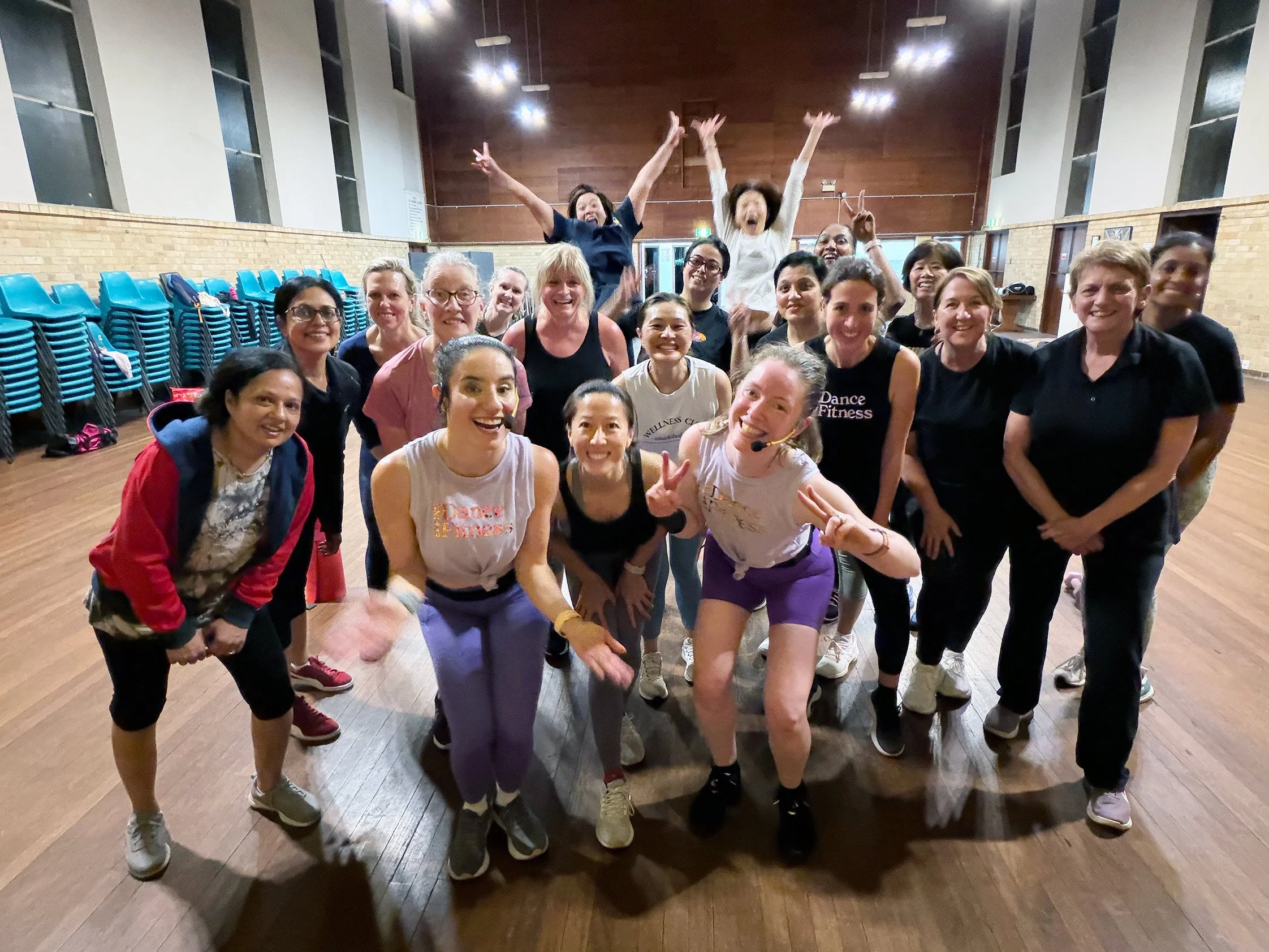 Group of women smiling and posing for a photo in a dance studio or gym, with some wearing fitness attire and a few wearing microphones.