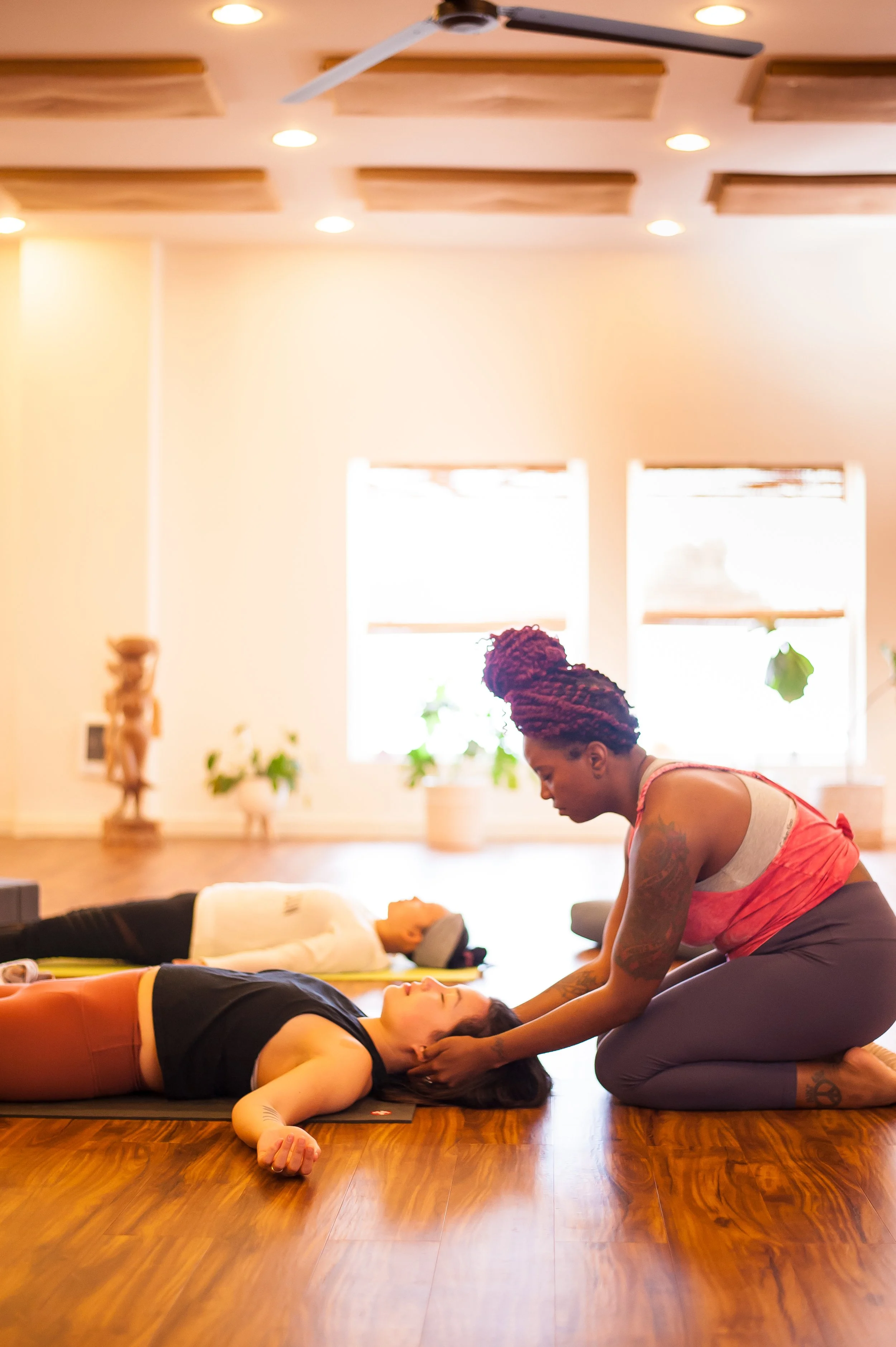 A yoga instructor gently assists a student lying on a yoga mat during a class, with another student in the background.