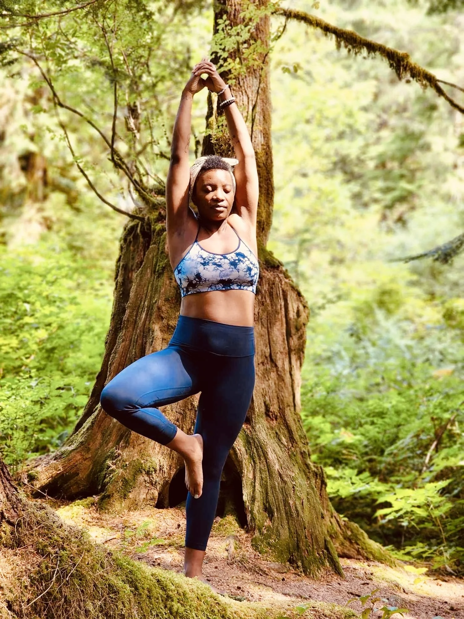 A woman practicing yoga in a forest, standing on one foot in tree pose with hands clasped above her head, surrounded by green trees and foliage.