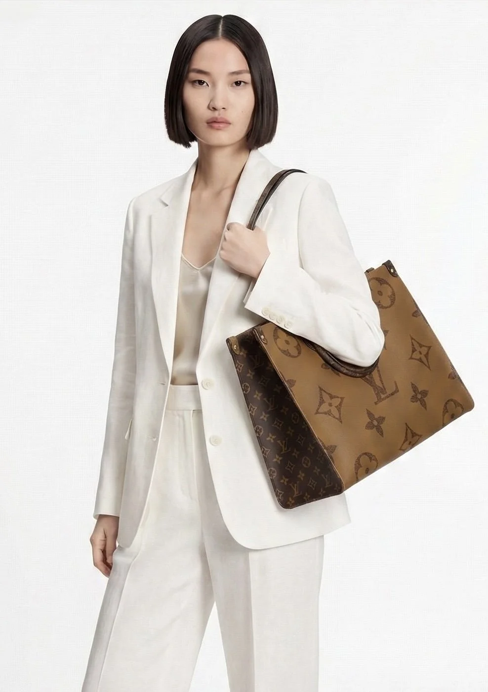 A woman with shoulder-length dark hair wearing a white suit with a blazer and matching pants, holding a large Louis Vuitton tote bag over her shoulder, against a plain white background.