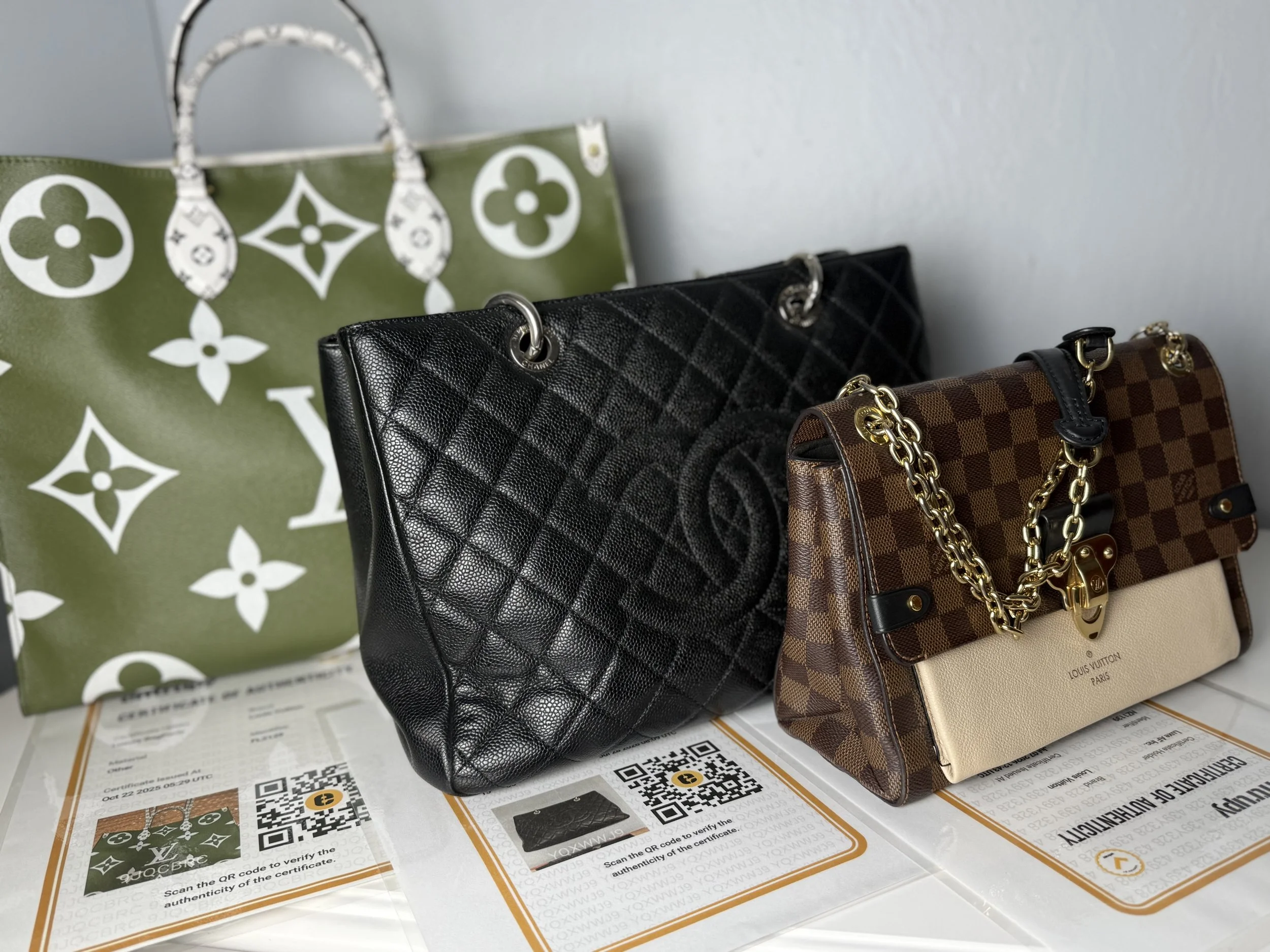 Three designer handbags (Louis Vuitton, Chanel, and Damier Ebene print Louis Vuitton) displayed on a table with printed documents and QR codes, with a large Louis Vuitton shopping bag in the background.