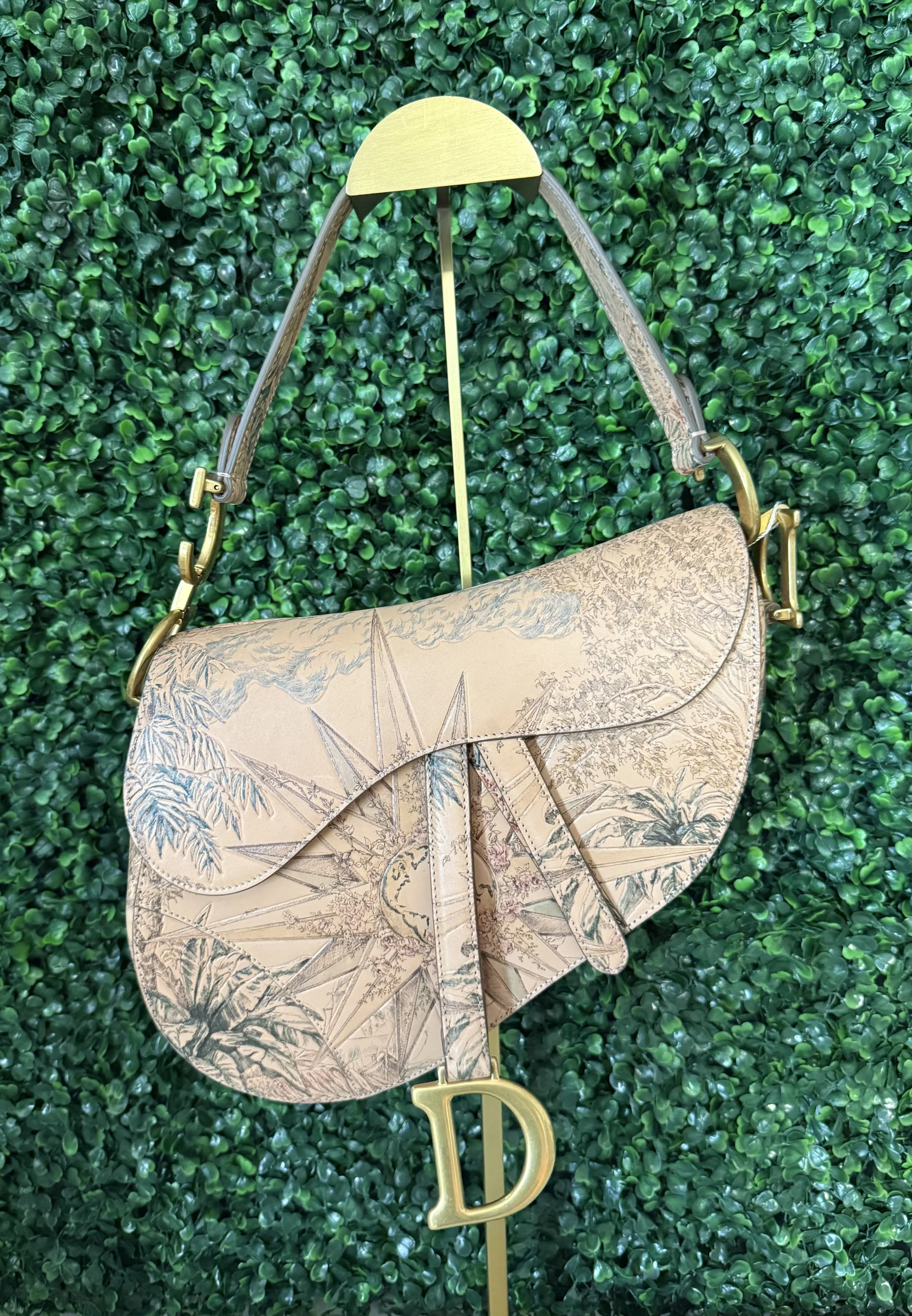 A designer handbag with a beige and brown map print design, hanging on a gold and gray stand against a green leafy background.