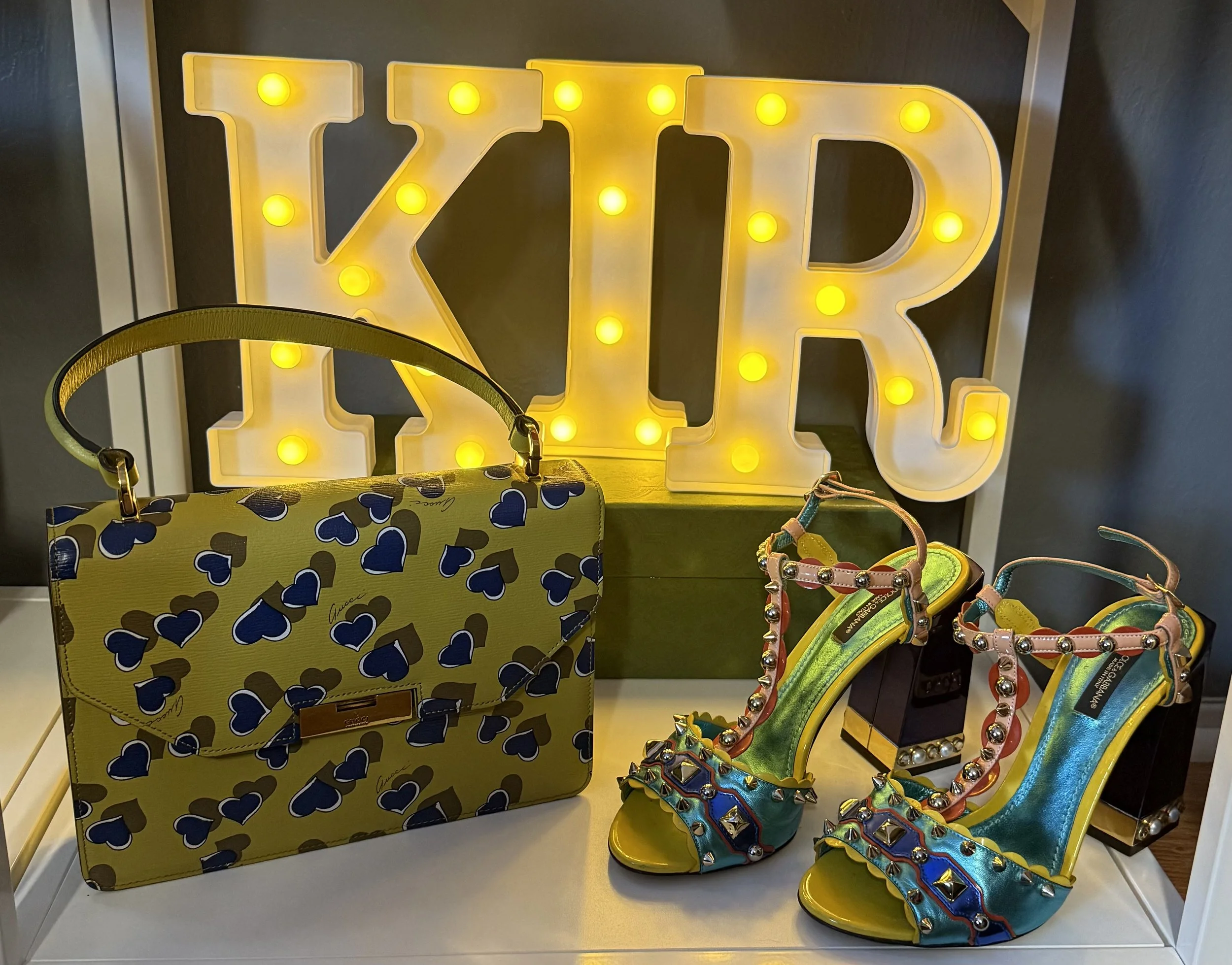 A display of colorful fashion accessories including a yellow handbag with blue and brown hearts, and a pair of high-heeled sandals with multicolored studs and embellishments, with a large illuminated sign spelling 'KIR' in the background.