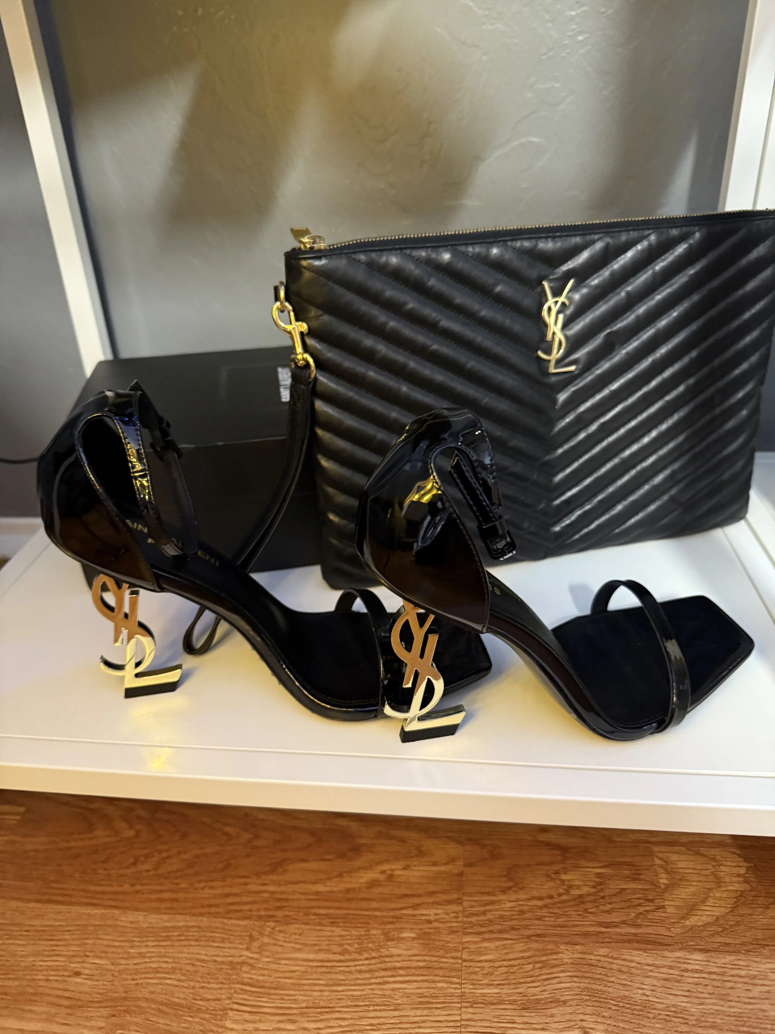 Black quilted YSL clutch bag and black high-heeled sandals with gold YSL logo accents on the heels, placed on a white surface.