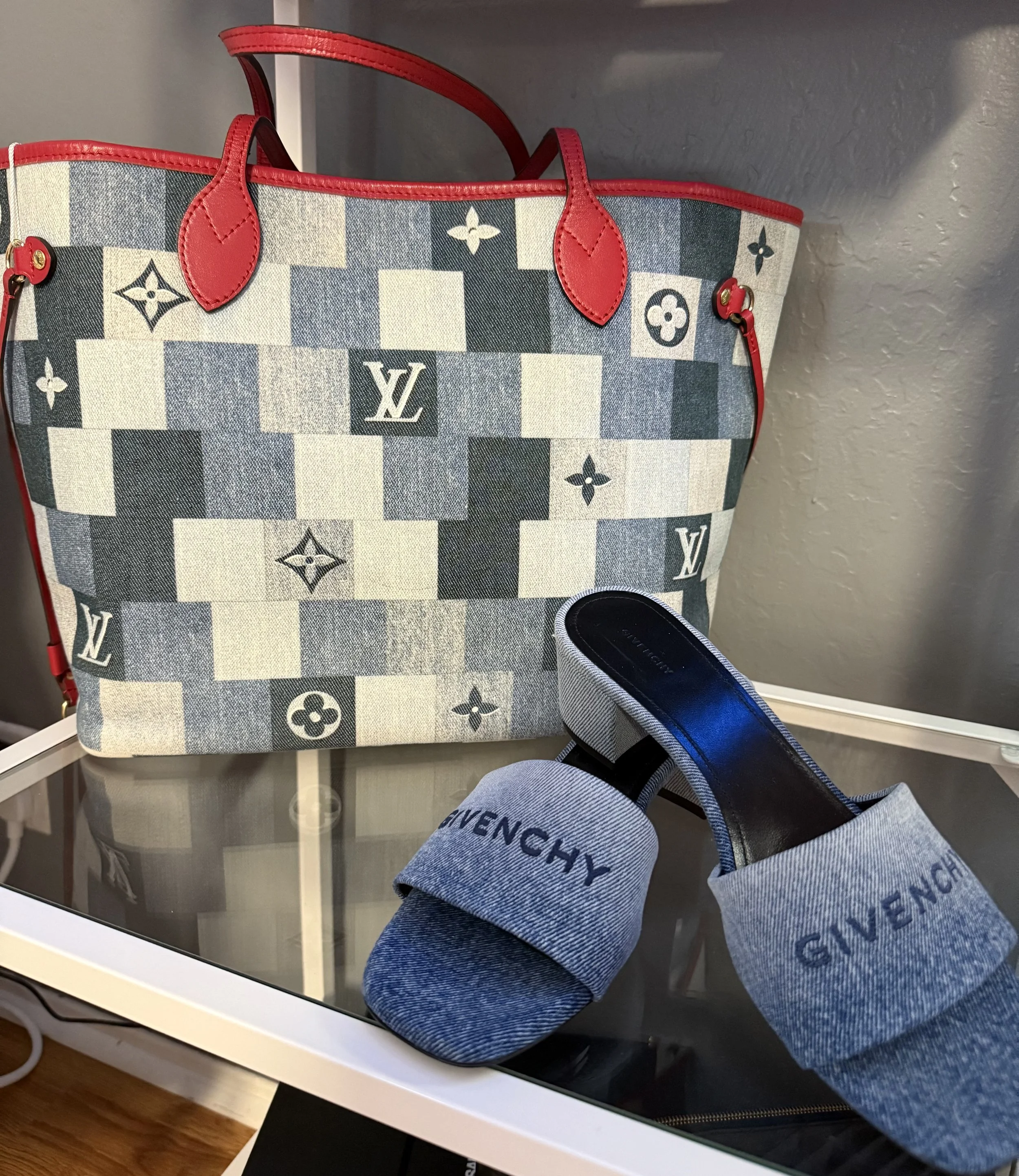 A Louis Vuitton tote bag with a checkered pattern and red leather handles, and a pair of denim Givenchy slide sandals with black soles placed in front of the bag, on a glass table.