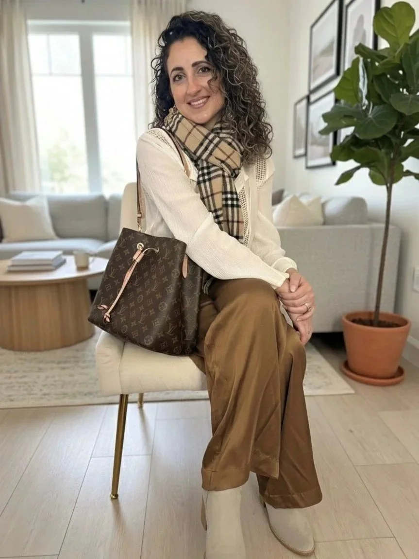 A woman with curly dark hair sitting on a cream-colored chair in a living room, smiling and wearing a beige sweater, plaid scarf, brown wide-leg pants, and beige boots. She has a Louis Vuitton handbag over her shoulder, and there is a large potted plant and artwork on the wall behind her.