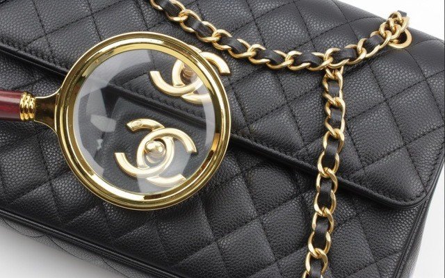 A black quilted leather Chanel purse with gold chain strap, a gold magnifying glass displaying the Chanel logo, and a black fabric tag.