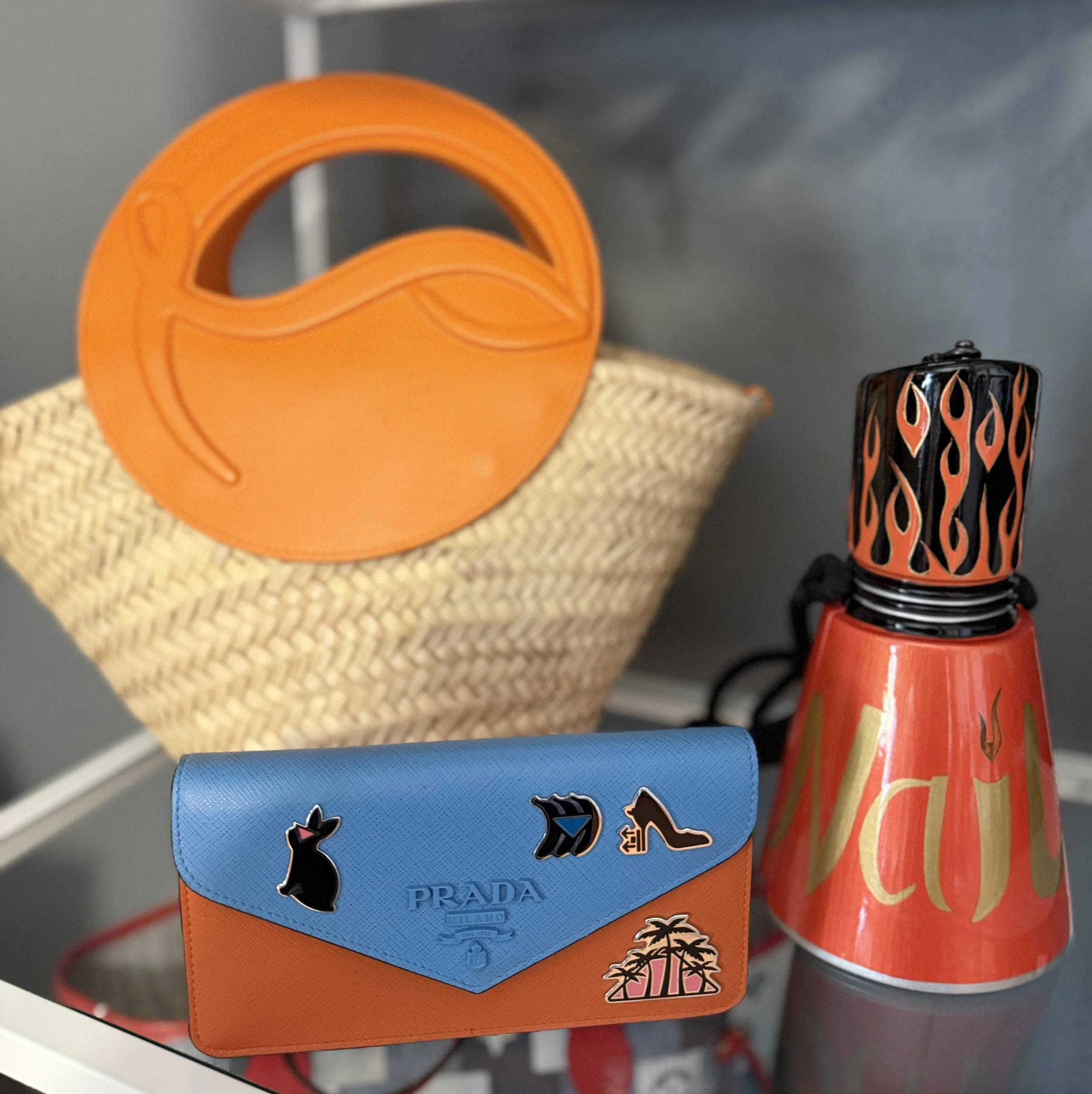 A tan woven basket with an orange circular plastic object on top, a red and black flame-patterned lighter, and a blue and orange Prada wallet with various pins, all placed on a white surface.