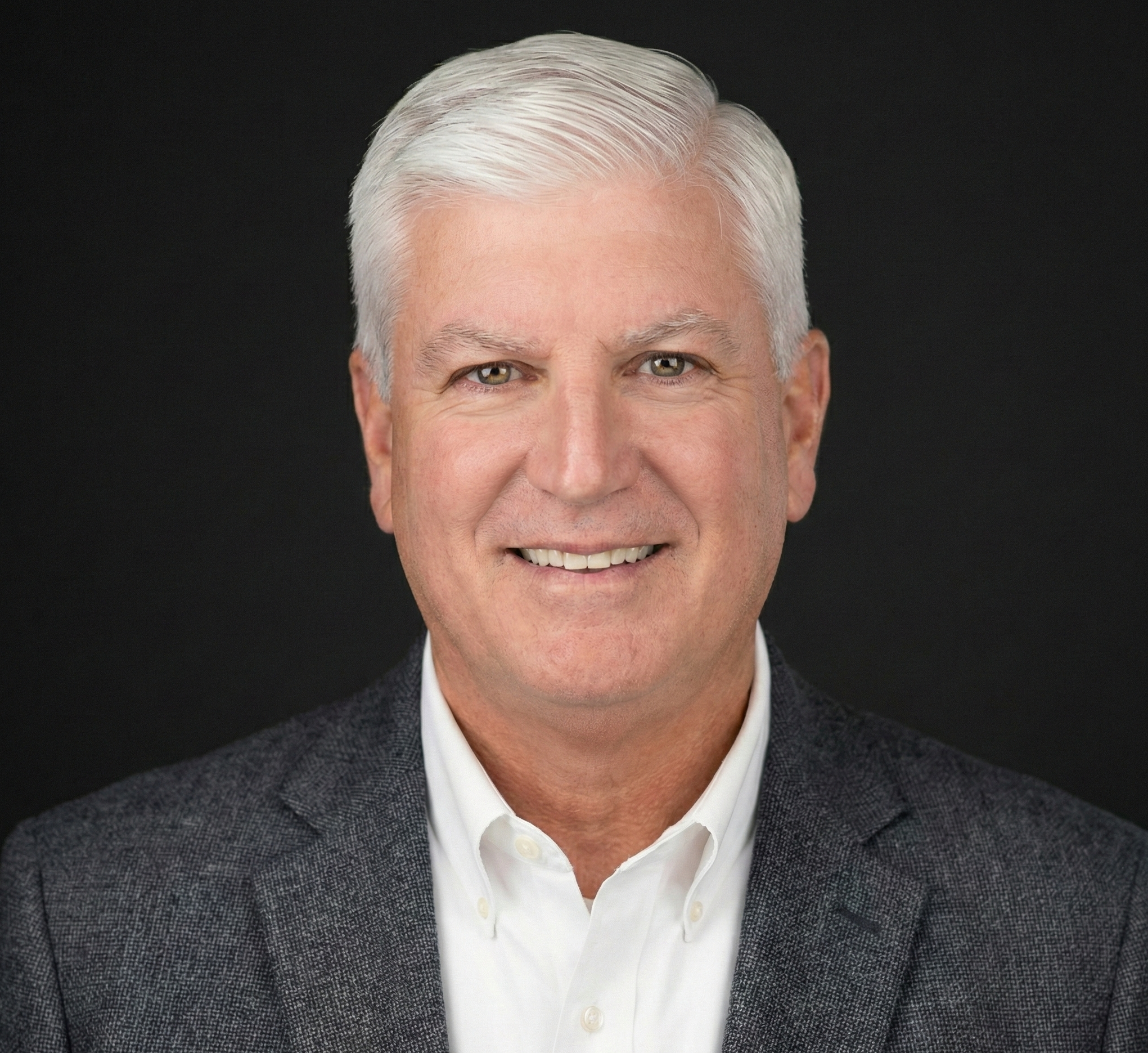Headshot of a middle-aged man with white hair, blue eyes, and a friendly expression, wearing a white collared shirt and a dark grey blazer, against a black background.