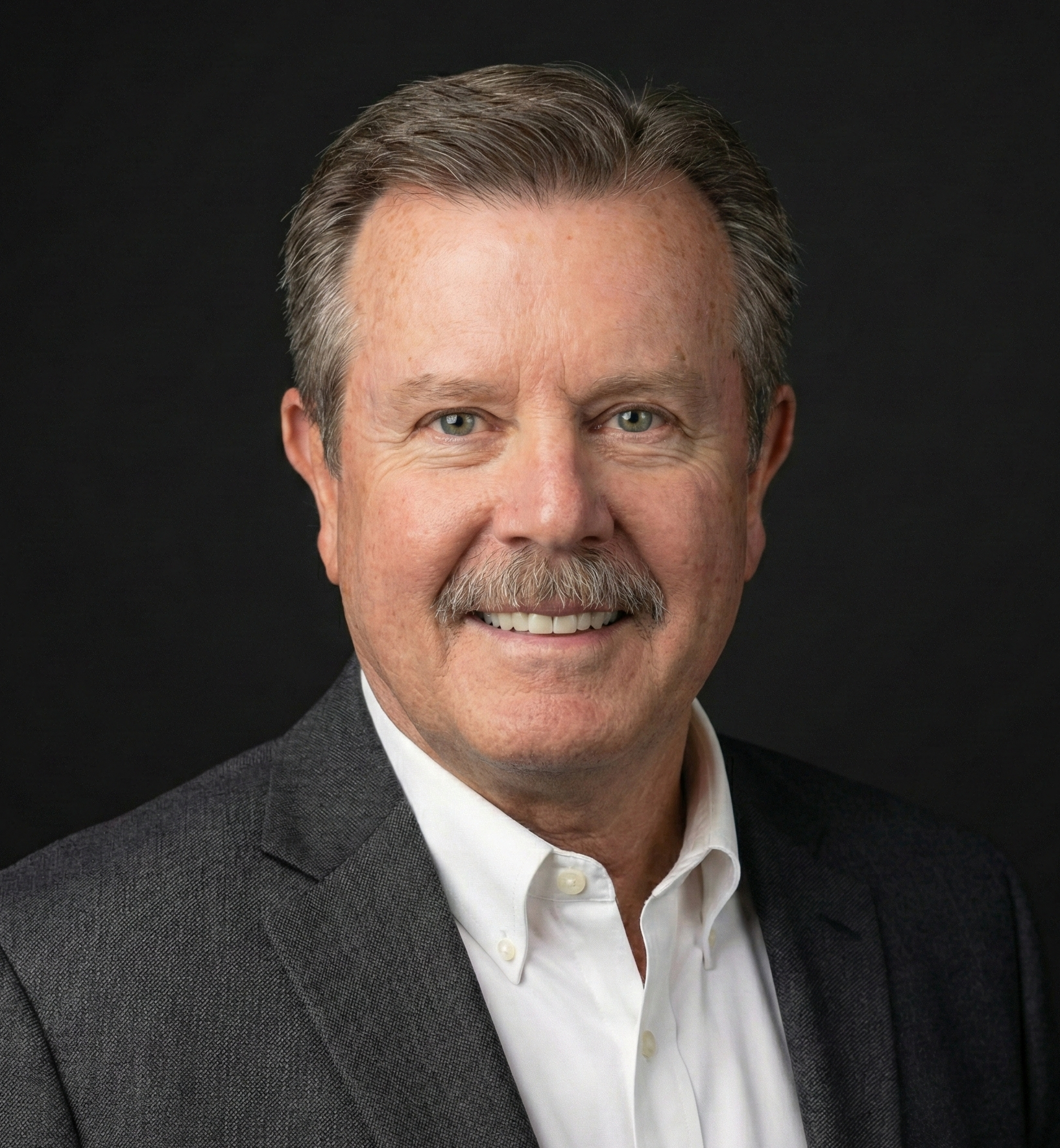 Portrait of a middle-aged man with gray hair, a mustache, blue eyes, wearing a dark gray suit jacket and a white dress shirt, smiling against a black background.